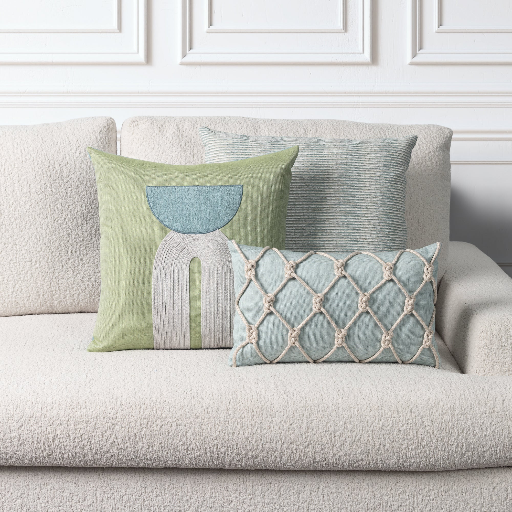 Three decorative throw pillows arranged on a textured light beige sofa against a white paneled wall. The pillows include a sage green one with a light blue and white abstract arch design, a pale blue-green ribbed square pillow, and a light blue rectangular pillow with cream rope-knotted diamond patterns.