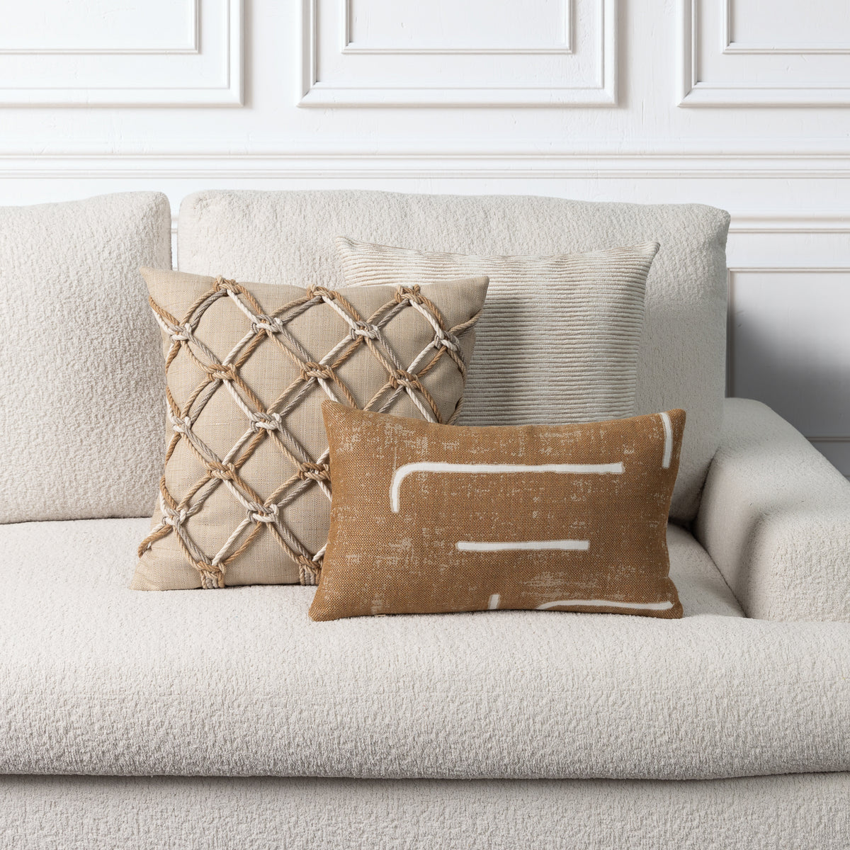 Three decorative throw pillows arranged on a light beige boucle sofa against a white paneled wall. The front pillow is tan with white abstract brushstroke lines, the center pillow is beige with a rope lattice pattern, and the back pillow is light beige with subtle vertical ribbing.