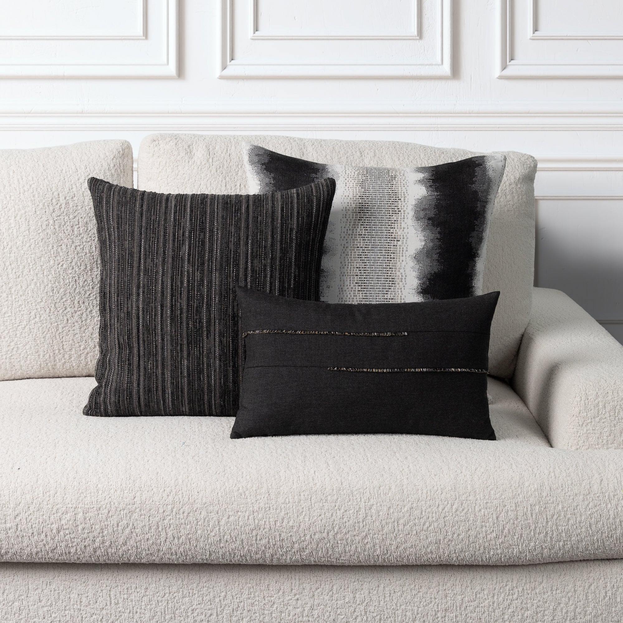 Three decorative throw pillows in black, gray, and white textured fabrics arranged on a light beige boucle sofa against a white paneled wall.