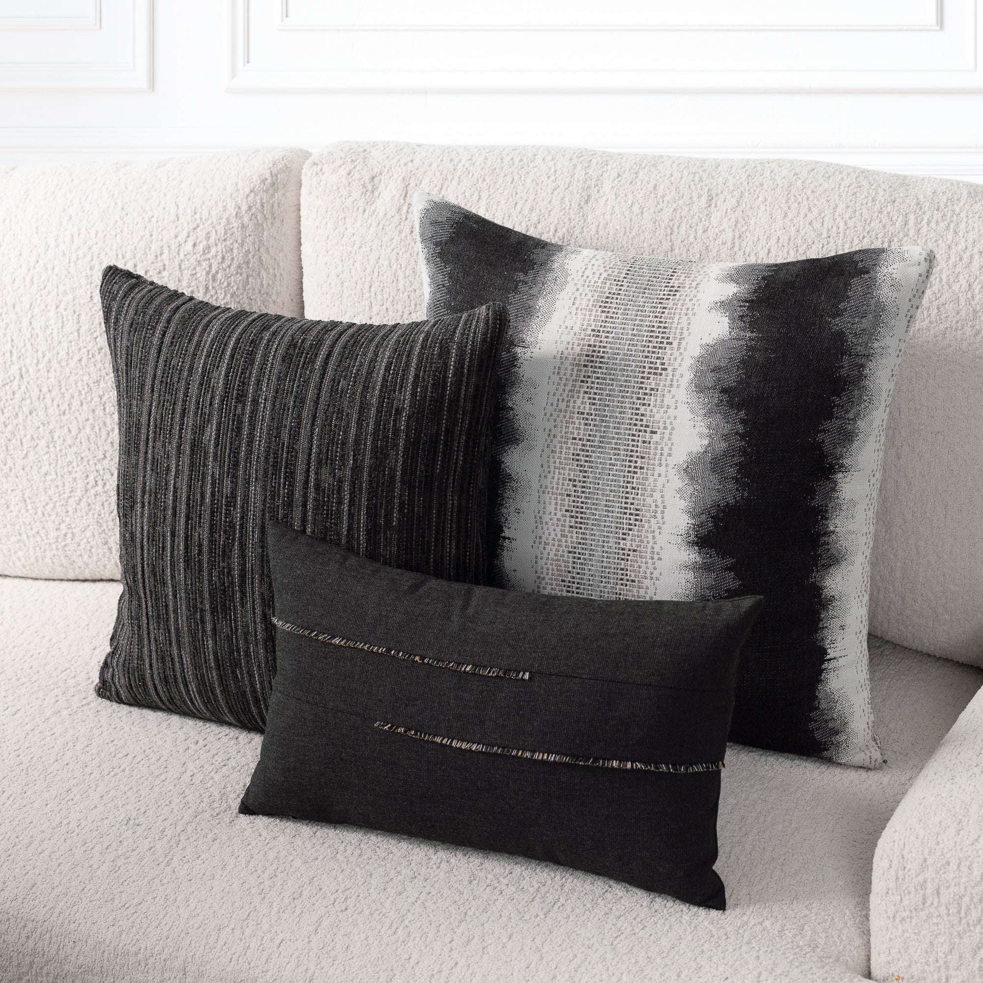 Three decorative throw pillows in black, gray, and white tones arranged on a textured cream sofa. One pillow has vertical gray stripes, one has a black and white brushstroke pattern, and the front pillow is black with a thin metallic horizontal stripe.