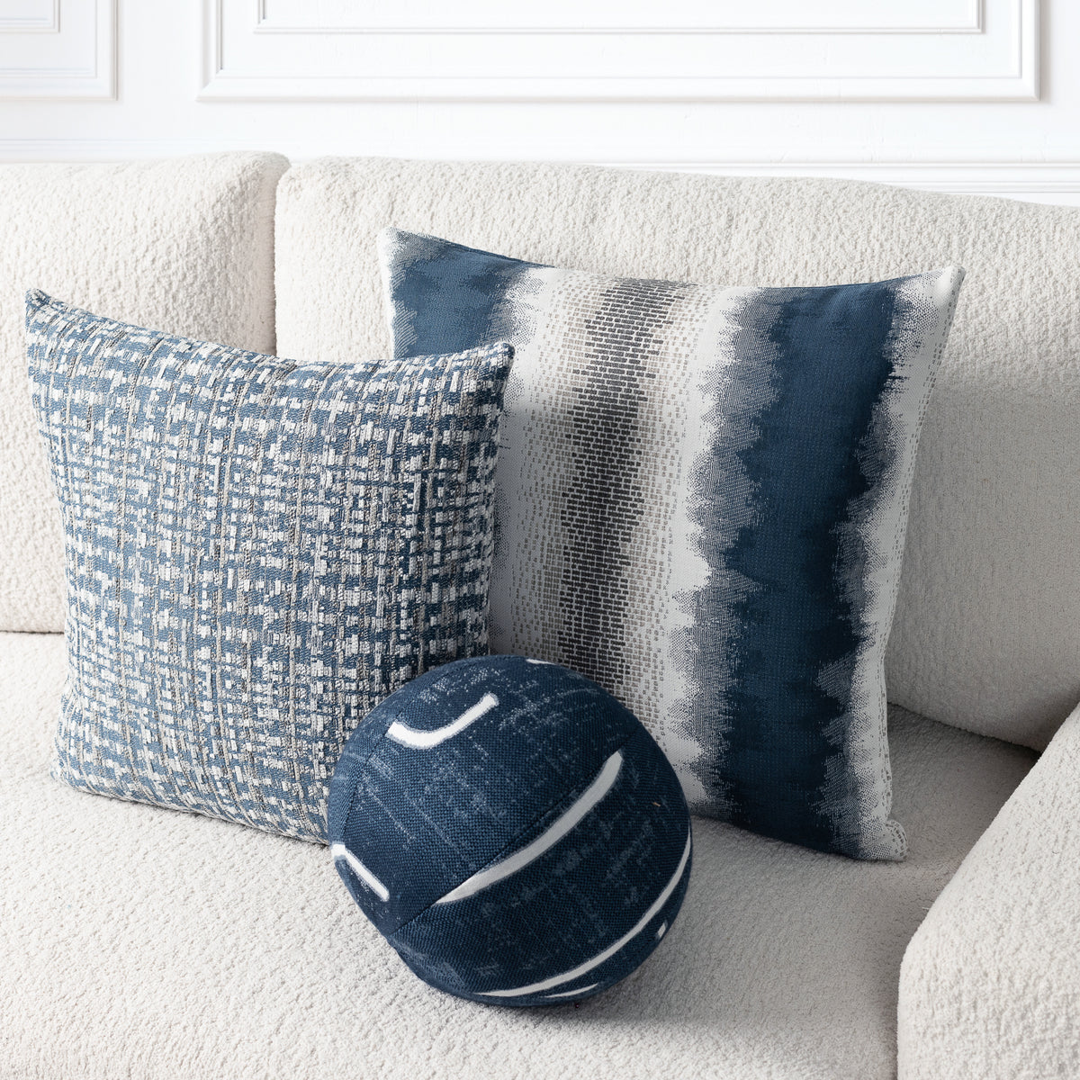Three decorative throw pillows arranged on a cream textured sofa: a square pillow with a blue-and-white woven pattern, a square pillow with vertical blue, gray, and white brushstroke stripes, and a round navy pillow with white curved stripes. White wall molding is visible in the background.