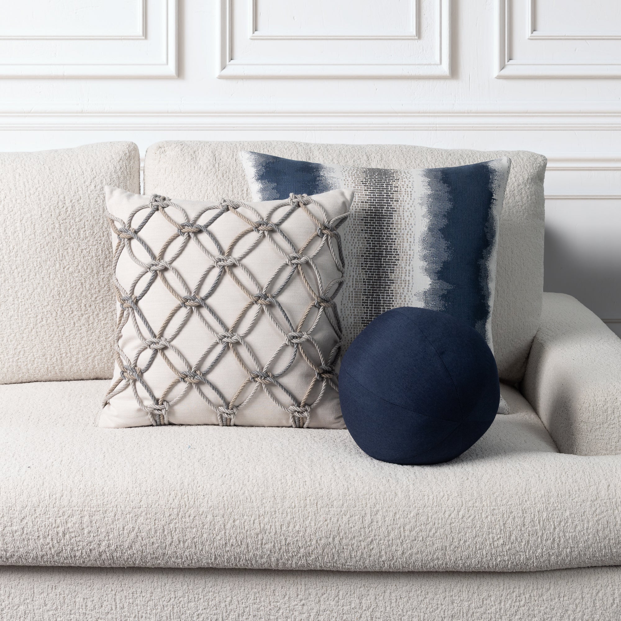 Three decorative throw pillows arranged on a light beige textured sofa against a white paneled wall: a square cream pillow with a gray rope lattice pattern, a vertical navy and gray striped pillow, and a round navy pillow.