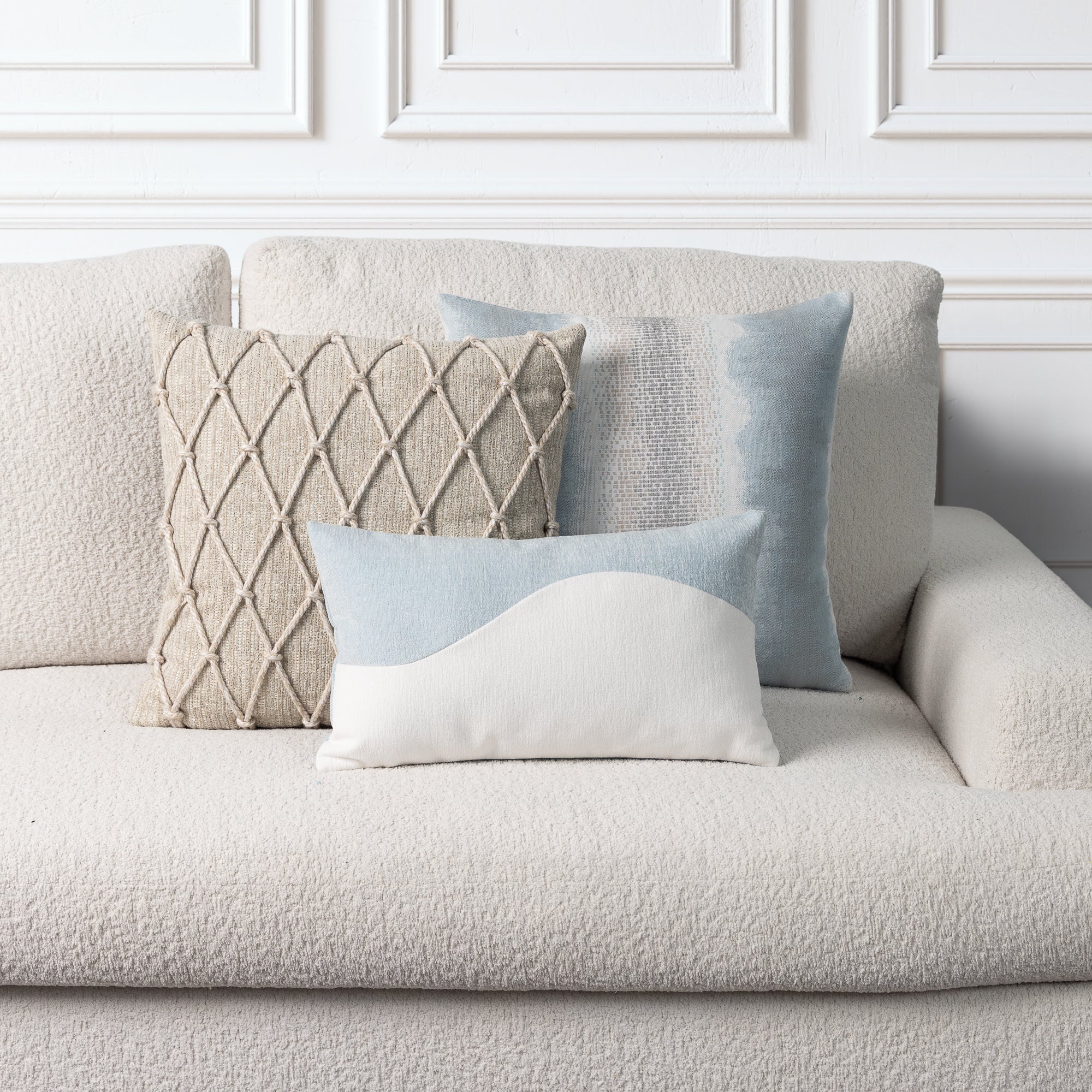 Three decorative throw pillows arranged on a textured cream boucle sofa against a white paneled wall. The pillows include a square beige woven diamond-pattern pillow, a light blue watercolor-style square pillow, a small white pillow with a light blue curved top, and a larger light blue rectangular pillow.