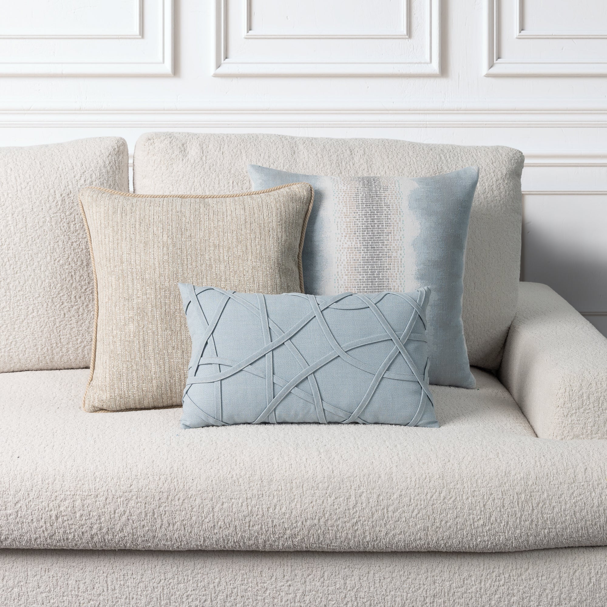 Three decorative throw pillows arranged on a textured cream boucle sofa against a white paneled wall. The set includes a large cream pillow, a light blue textured pillow, and a smaller light blue lumbar pillow with intersecting stitched lines.