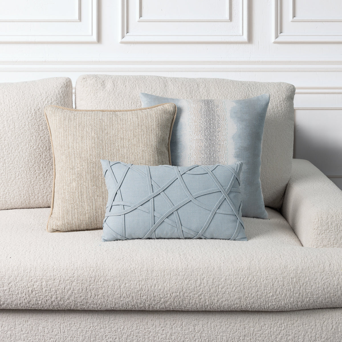 Three decorative throw pillows arranged on a textured cream boucle sofa against a white paneled wall. The set includes a large cream pillow, a light blue textured pillow, and a smaller light blue lumbar pillow with intersecting stitched lines.