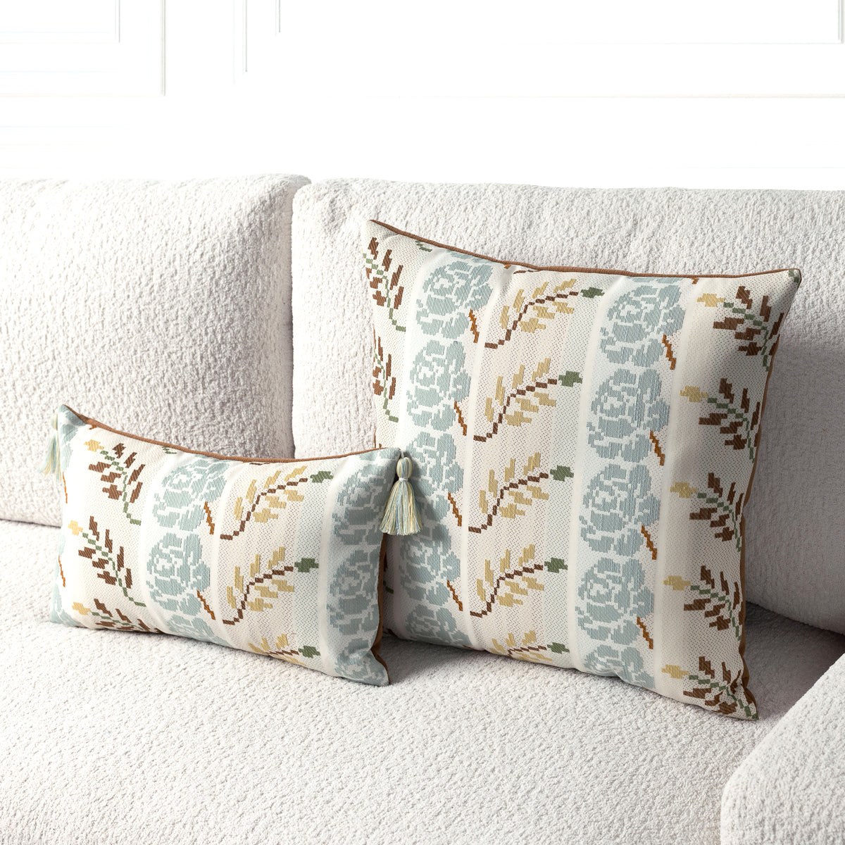 Two decorative throw pillows with a floral pattern in muted blue-gray, olive green, and brown on a cream background, styled on a textured light beige sofa.