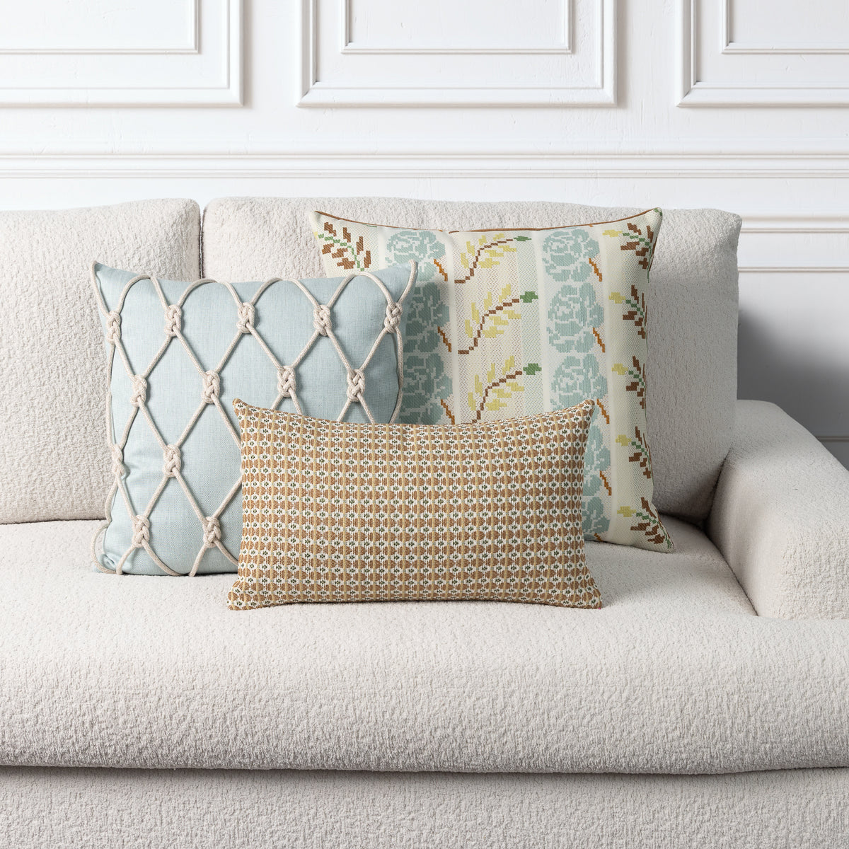 Three decorative throw pillows arranged on a light beige textured sofa against a white paneled wall. The front pillow is rectangular with a gold, brown, and beige geometric pattern.