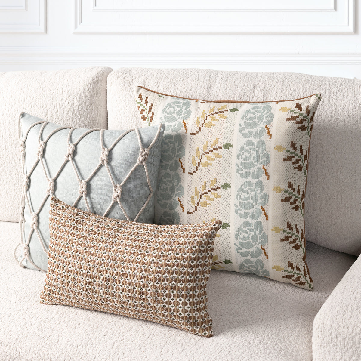 Three decorative throw pillows arranged on a cream boucle sofa against a white paneled wall. The pillows include a sage green one with a knotted diamond pattern, a cream one with a muted floral and leaf print in sage green, mustard, and brown, and a smaller rectangular tan one with a geometric woven pattern.