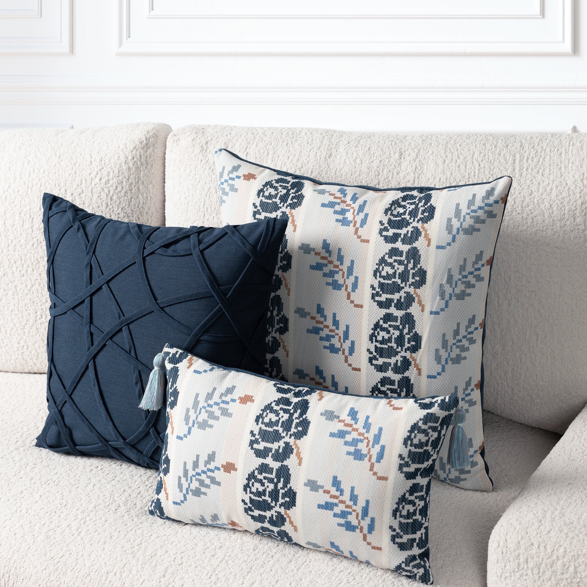 Three decorative throw pillows arranged on a cream boucle sofa. One pillow is dark navy blue with a crisscross textured pattern, and two other pillows feature a cream background with navy floral motifs and blue leaf accents.