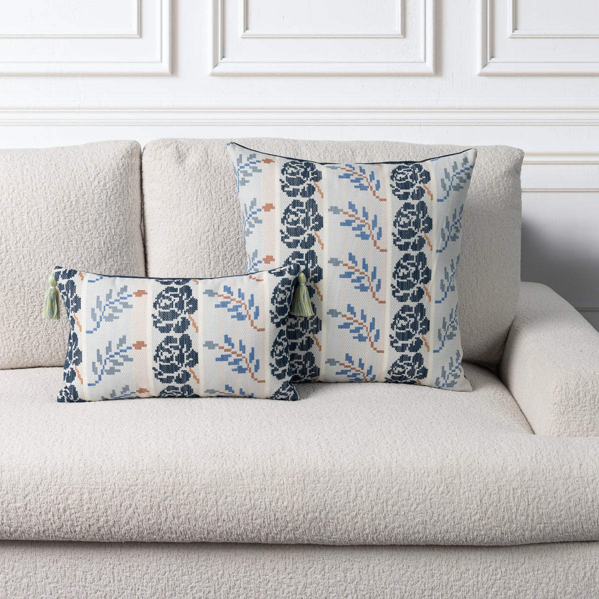 Two decorative throw pillows with a navy blue and light blue floral pattern on a cream background, styled on a textured beige sofa against a white paneled wall.