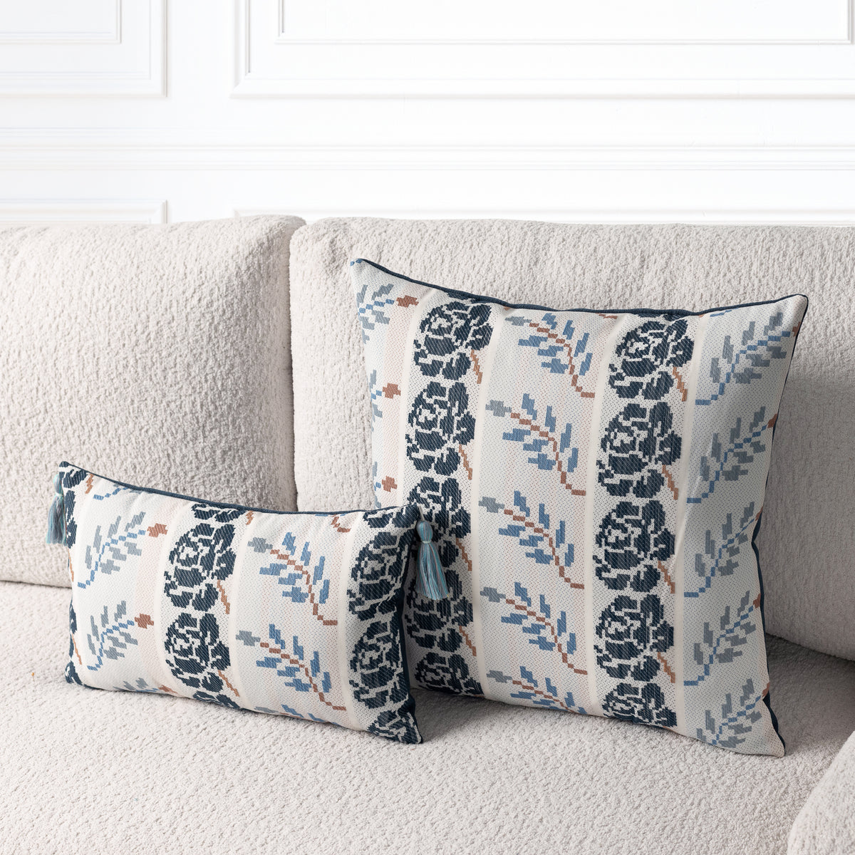 Two decorative throw pillows with a navy blue floral and leaf pattern on a cream background, styled on a textured beige sofa.
