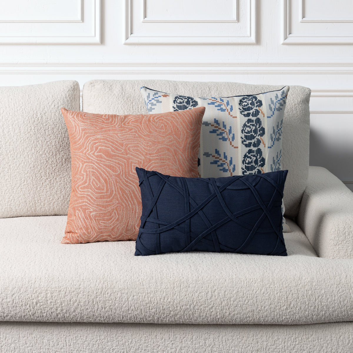 Three decorative throw pillows arranged on a textured cream boucle sofa against a white paneled wall. The front pillow is dark navy blue with a crisscross stitched pattern, the middle pillow is a muted peach with a wavy line pattern, and the back pillow has a navy floral pattern on a light background.