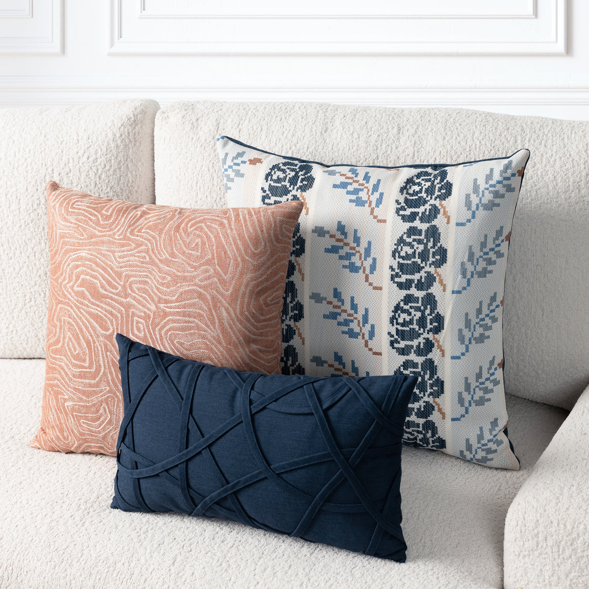 Three decorative throw pillows arranged on a cream textured sofa: a peach pillow with a white abstract line pattern, a navy pillow with a crisscross quilted texture, and a white pillow with a navy floral and blue leaf pattern.