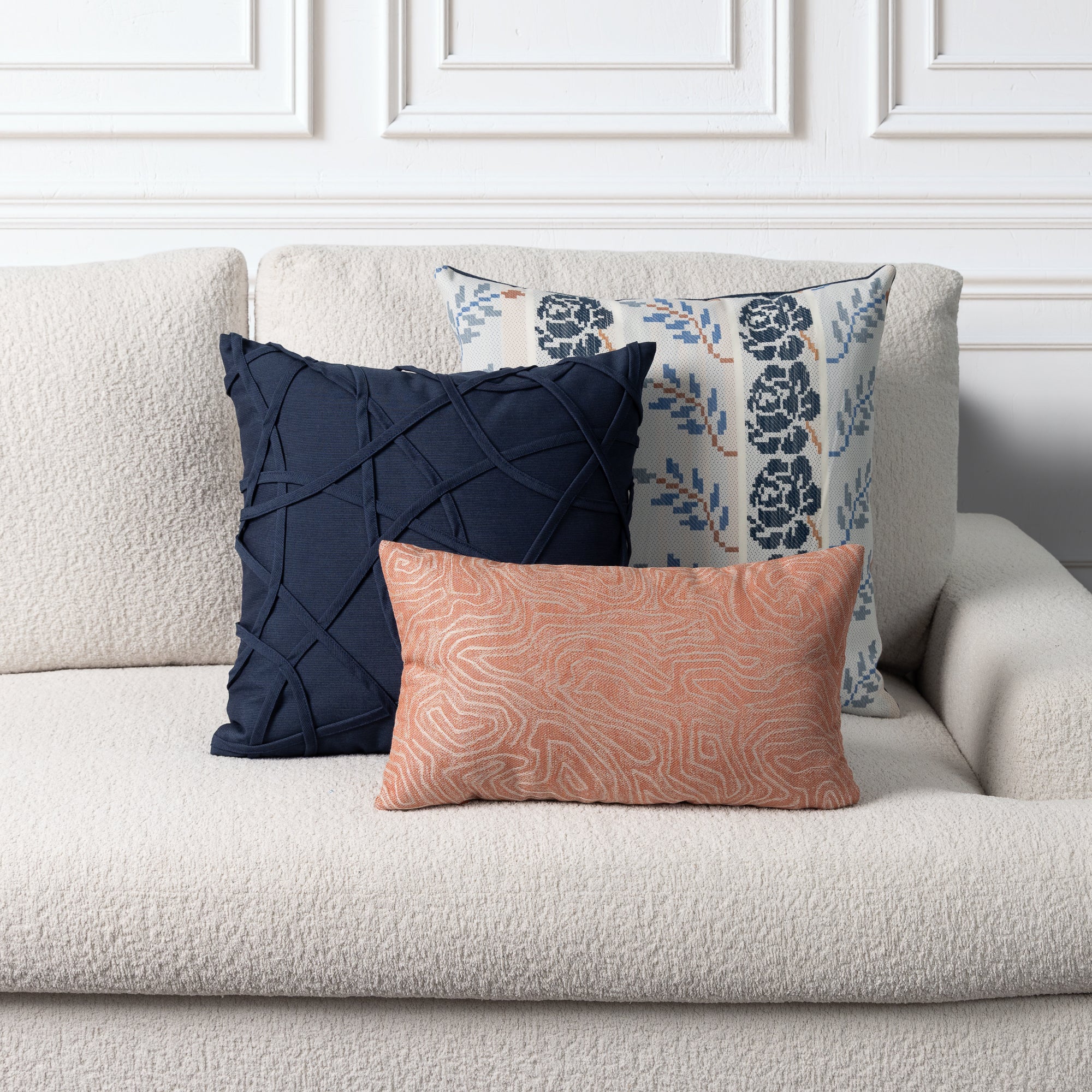 Three decorative throw pillows arranged on a light beige boucle sofa against a white paneled wall. The front pillow is a peachy pink with a wavy pattern, the back-left pillow is dark navy with a quilted texture, and the back-right pillow is white with a navy floral pattern.