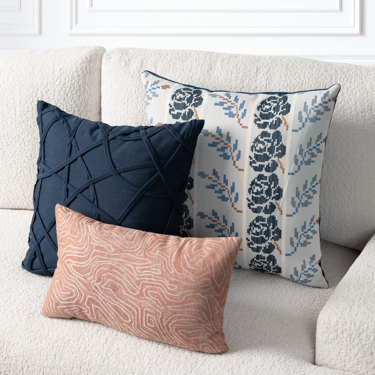 Three decorative throw pillows arranged on a textured cream sofa: a dark navy pillow with a quilted geometric pattern, a square cream pillow with a blue floral ikat pattern, and a rectangular peach pillow with a wavy abstract line pattern.