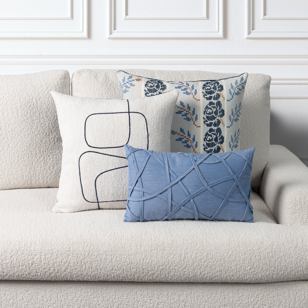Three decorative throw pillows arranged on a textured cream sofa against a white paneled wall. The front pillow is light blue with a crisscross stitched pattern, the middle pillow is cream with dark blue abstract line art, and the back pillow is cream with blue and gray floral embroidery.