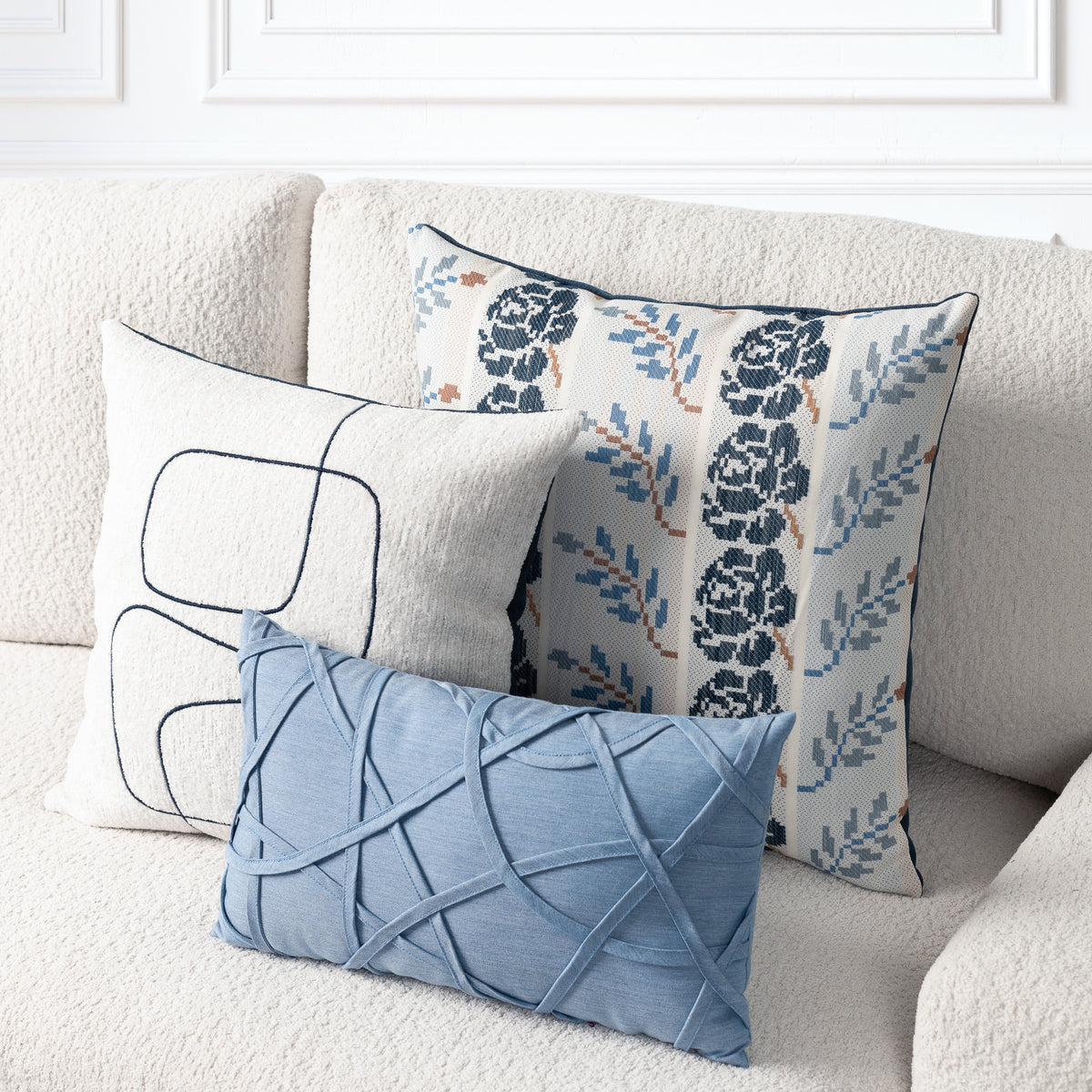 Three decorative throw pillows arranged on a cream boucle sofa: a light blue rectangular pillow with an abstract crisscross texture, a white pillow with dark navy line art, and a cream pillow with a navy floral and blue leaf pattern.