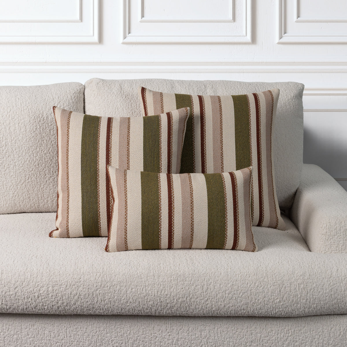 Three striped throw pillows in beige, brown, and olive green tones arranged on a light beige textured sofa against a white paneled wall.