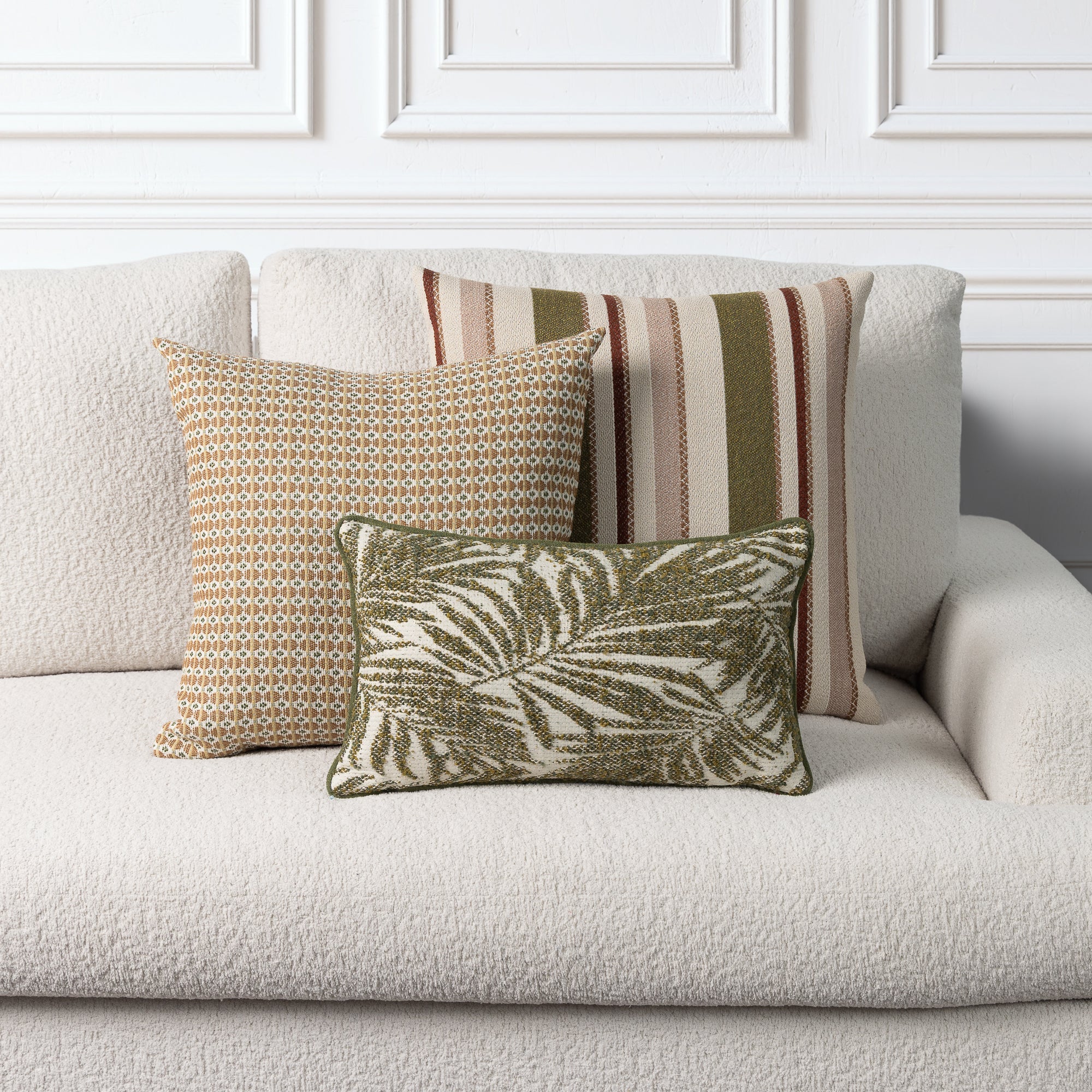 Three decorative throw pillows arranged on a light beige boucle sofa against a white paneled wall. The front pillow features an olive green and cream palm leaf pattern, the center pillow has a tan and cream geometric design, and the back pillow shows vertical stripes in beige, rust, and olive green.