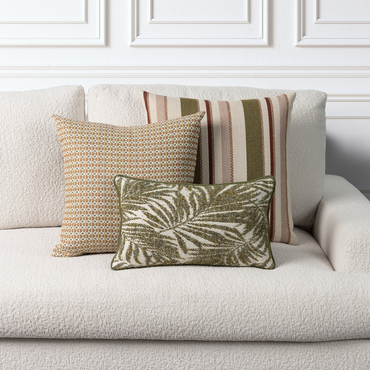 Three decorative throw pillows arranged on a light beige boucle sofa against a white paneled wall. The front pillow features an olive green and cream palm leaf pattern, the center pillow has a tan and cream geometric design, and the back pillow shows vertical stripes in beige, rust, and olive green.