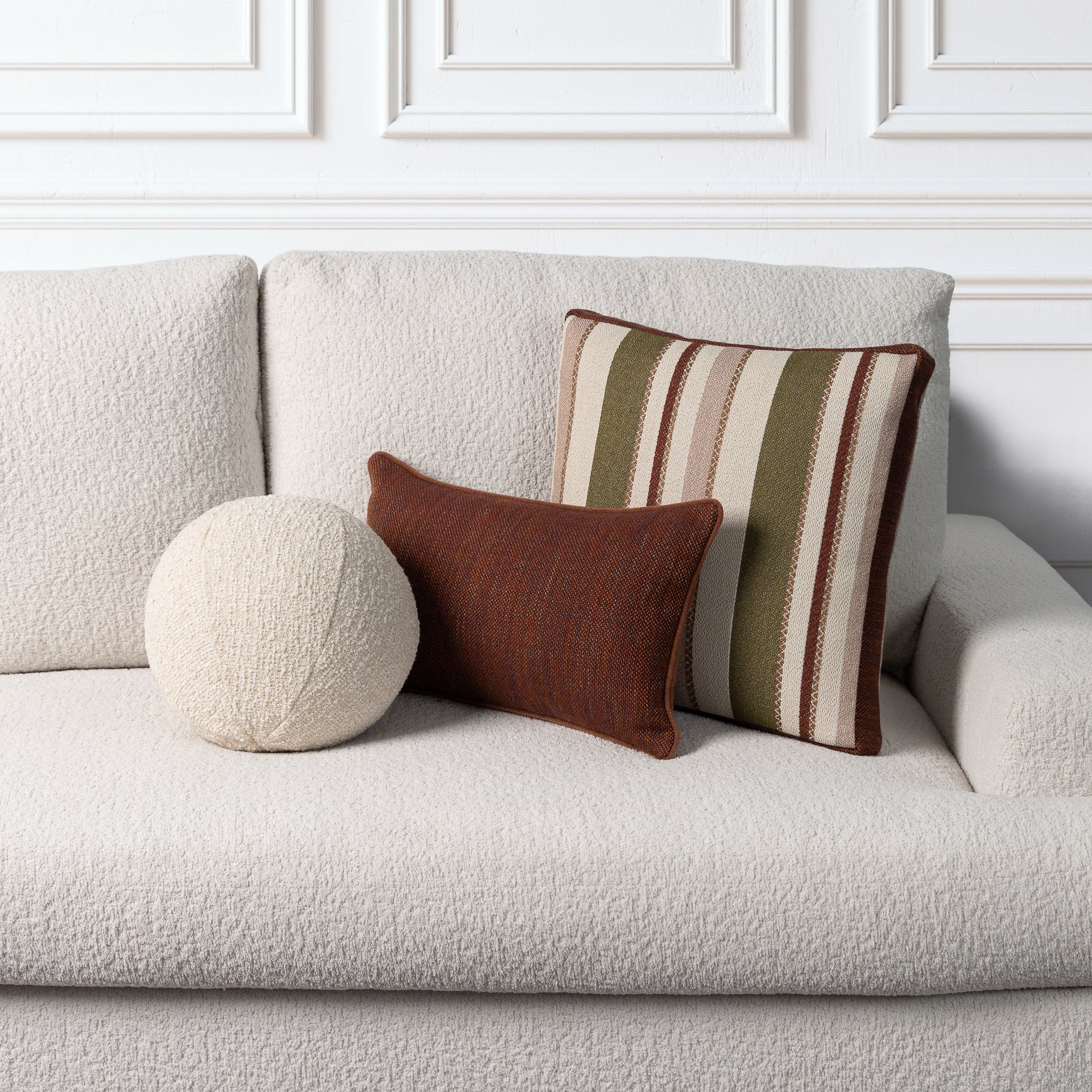 Three decorative throw pillows arranged on a light beige bouclé sofa against a white paneled wall. The set includes a round cream pillow, a small rust-brown rectangular pillow, and a larger square striped pillow in cream, olive green, and brown.
