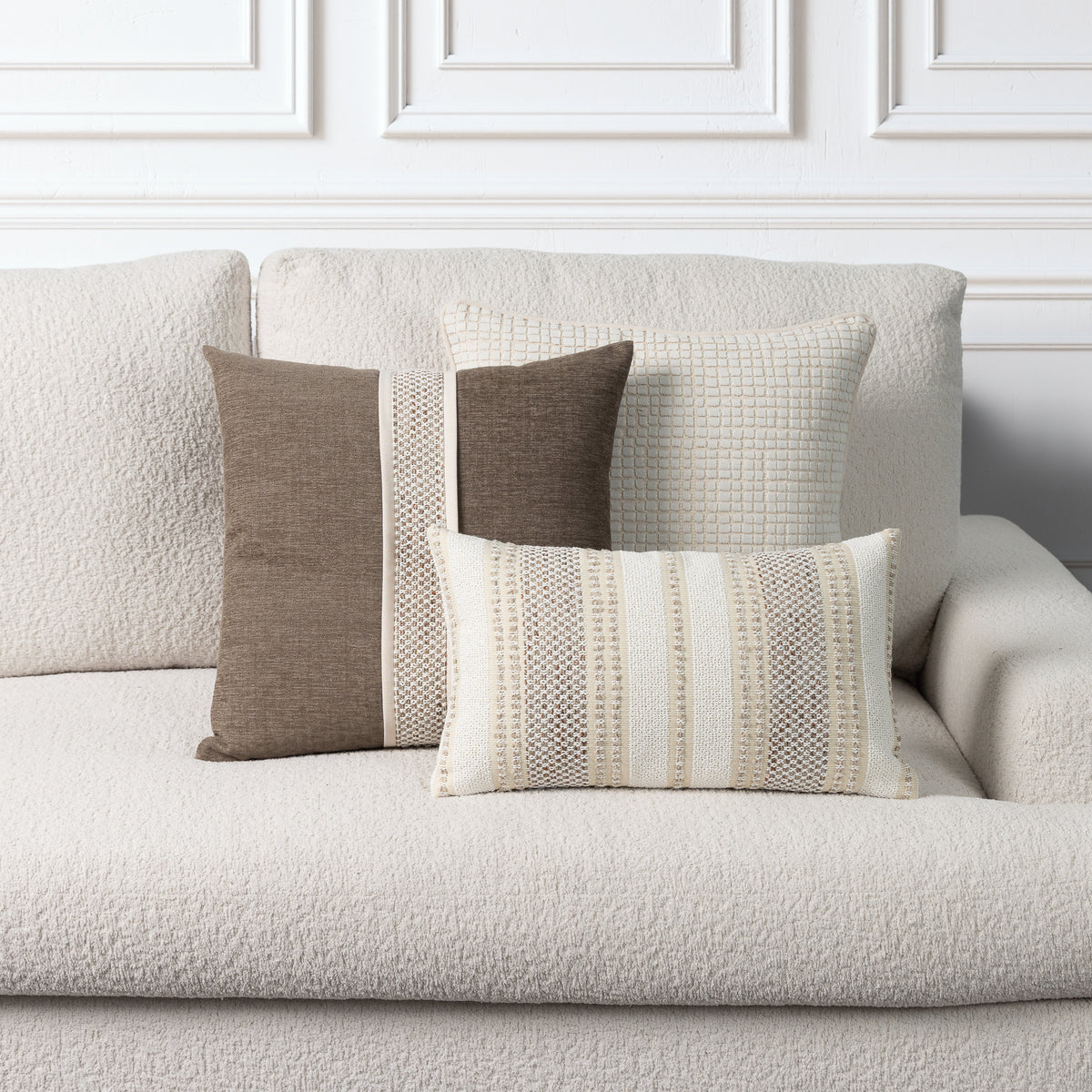 A set of textured throw pillows in neutral tones of beige, cream, and taupe arranged on a light beige boucle sofa against a white paneled wall.