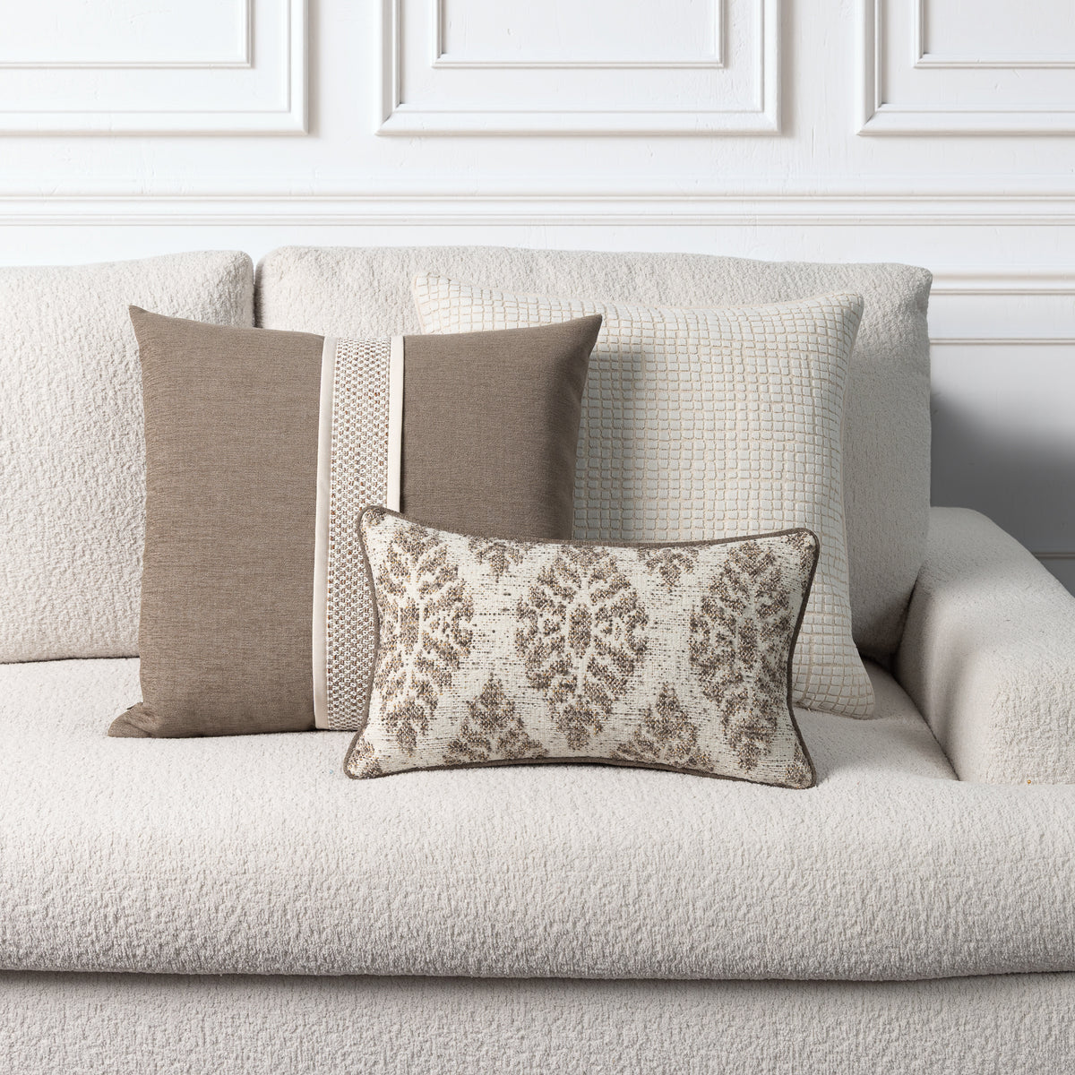 Three decorative throw pillows in neutral beige and brown tones arranged on a textured cream sofa against a white paneled wall.