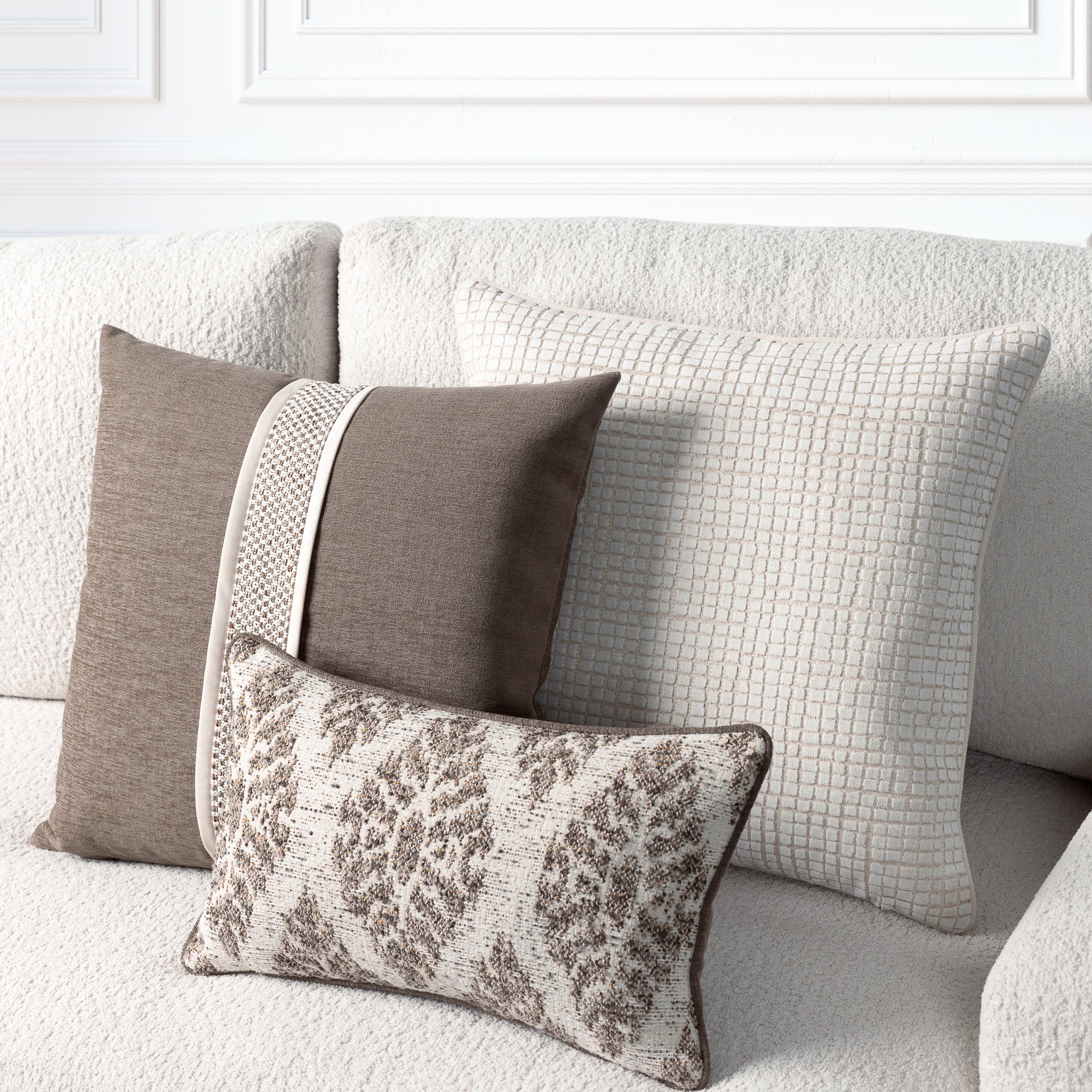 Three decorative throw pillows in neutral tones arranged on a textured light beige sofa. The set includes a solid taupe pillow, a cream grid-patterned pillow, and a smaller patterned lumbar pillow with taupe and cream damask-style motifs.
