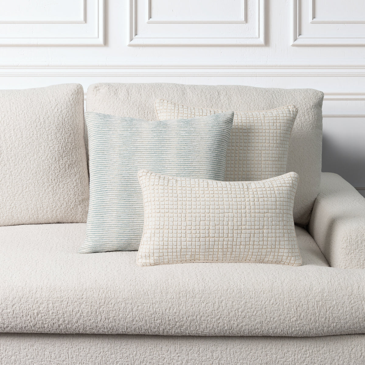Three textured throw pillows in neutral tones arranged on a light beige boucle sofa against a white paneled wall.