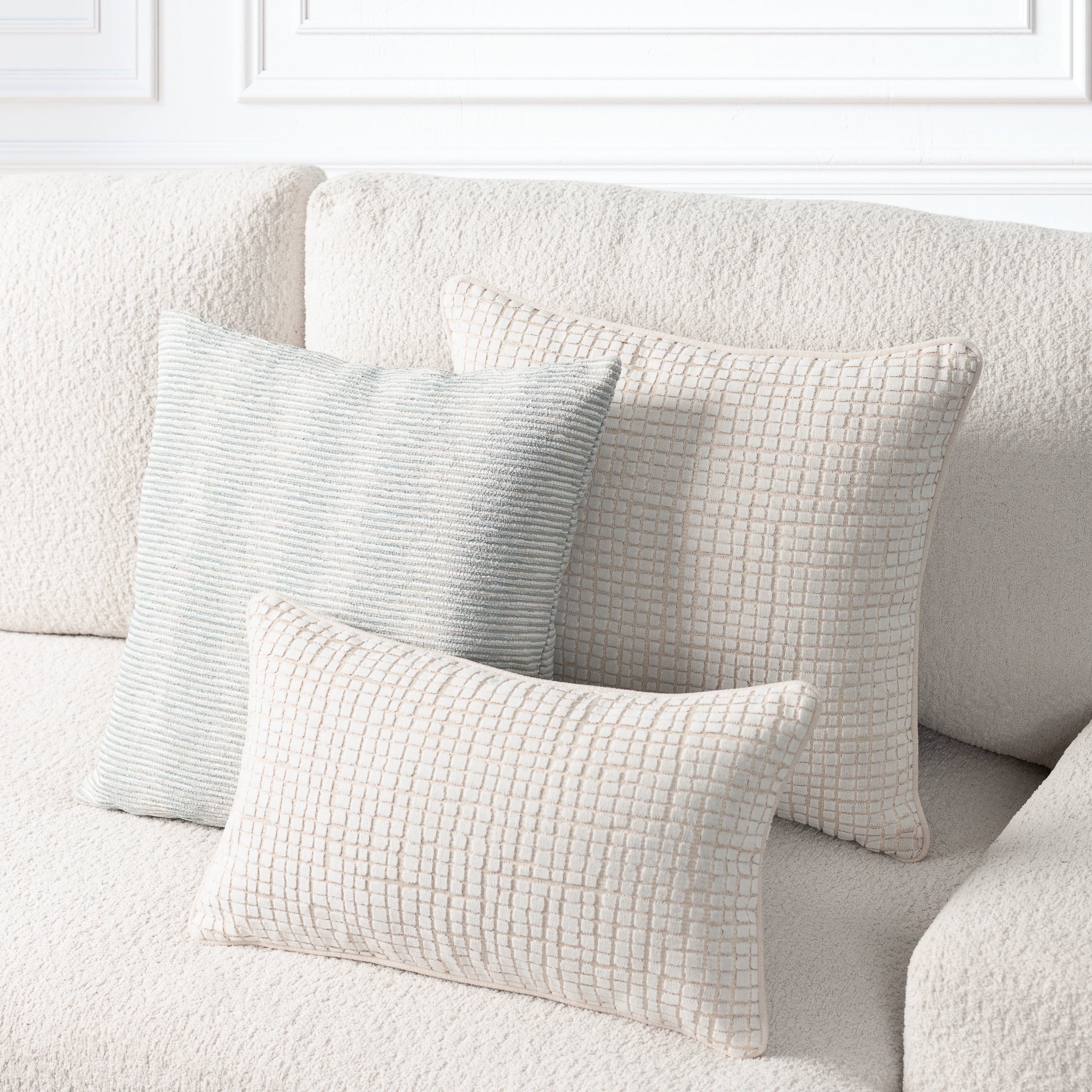 Three textured throw pillows in neutral tones arranged on a cream boucle sofa against a white paneled wall.
