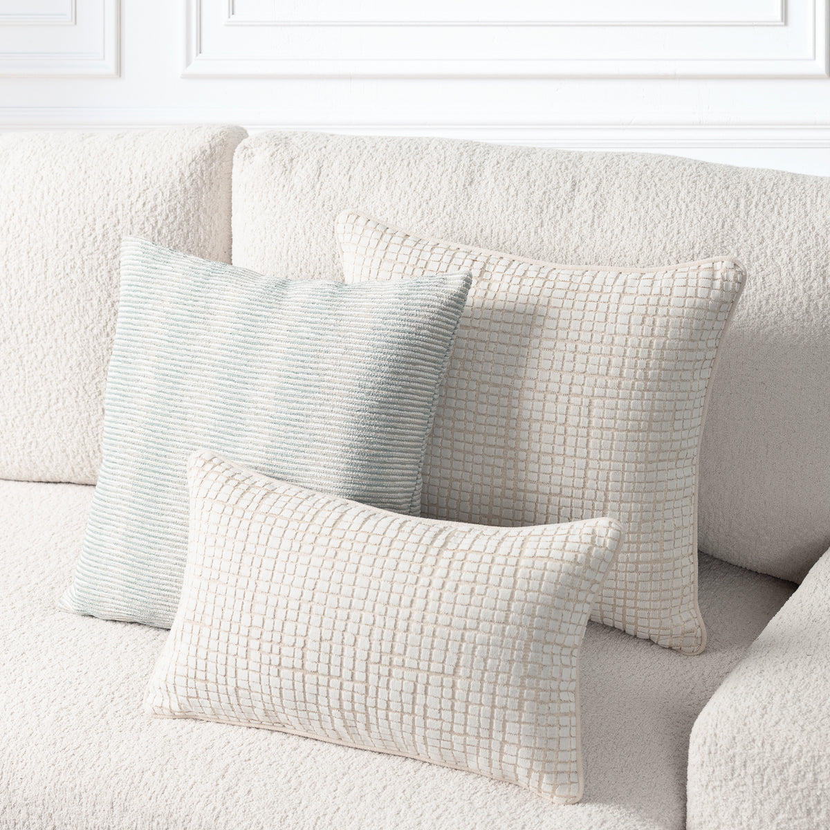 Three textured throw pillows in neutral tones arranged on a cream boucle sofa against a white paneled wall.
