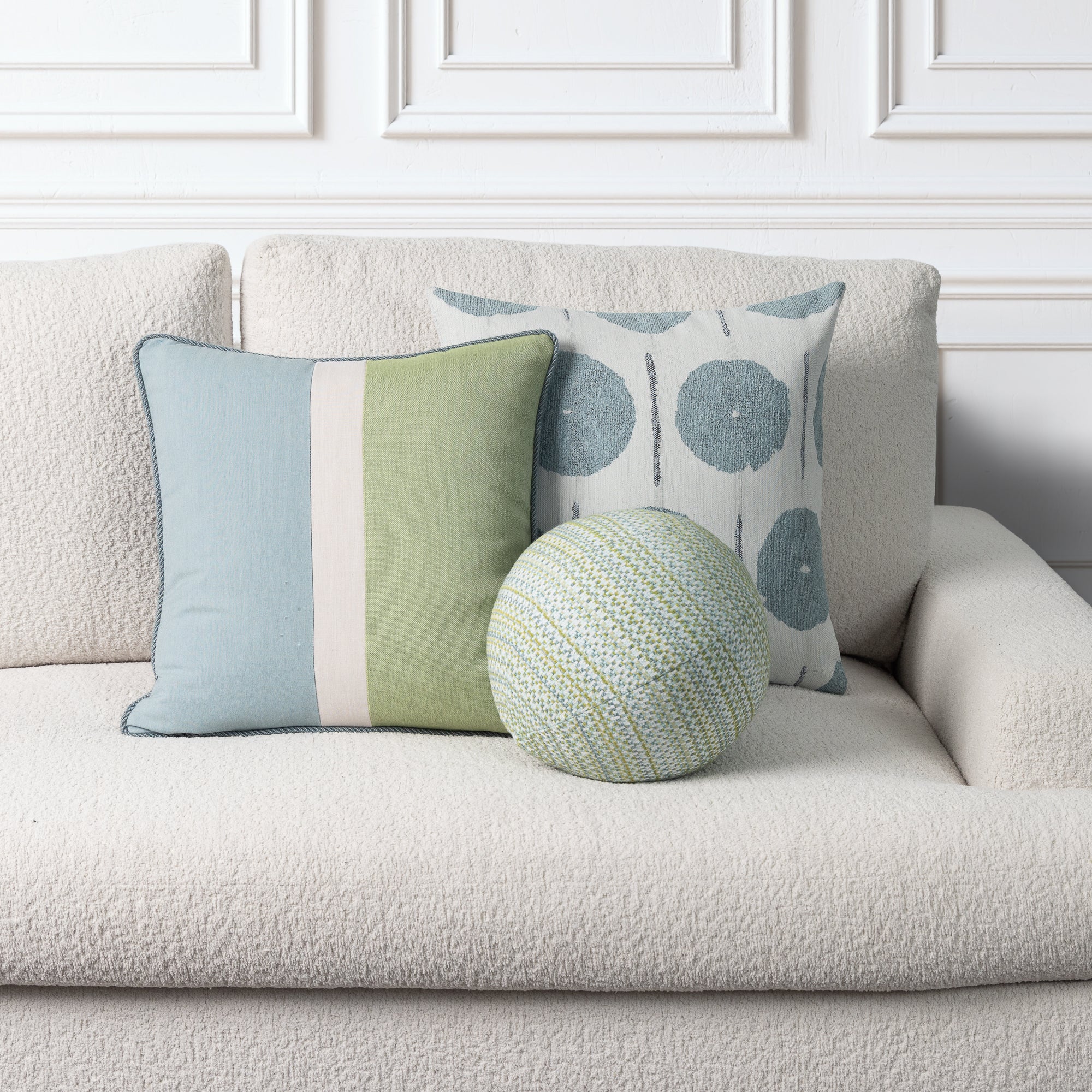 Three decorative throw pillows arranged on a textured cream boucle sofa against a white paneled wall. One square pillow is light blue and sage green with a white stripe; one square pillow has a white background with blue-gray circular motifs; one round pillow is green, blue, and white patterned.