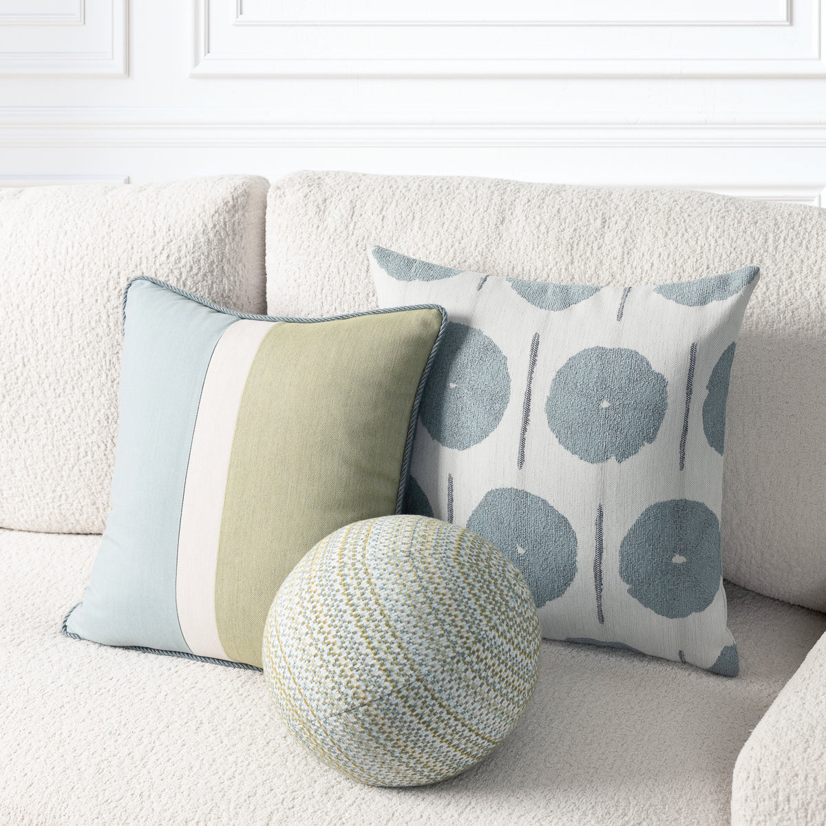 Three decorative throw pillows arranged on a textured cream sofa: a square pillow with vertical sage green, cream, and pale blue stripes, a square pillow with blue-gray circular motifs on a light background, and a round woven pouf in front.
