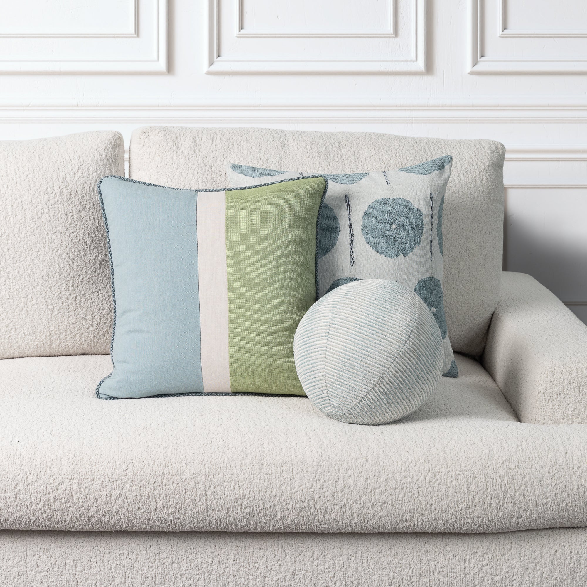 Three decorative throw pillows arranged on a textured cream boucle sofa against a white paneled wall. The front-left pillow is square with light blue, sage green, and off-white vertical stripes.