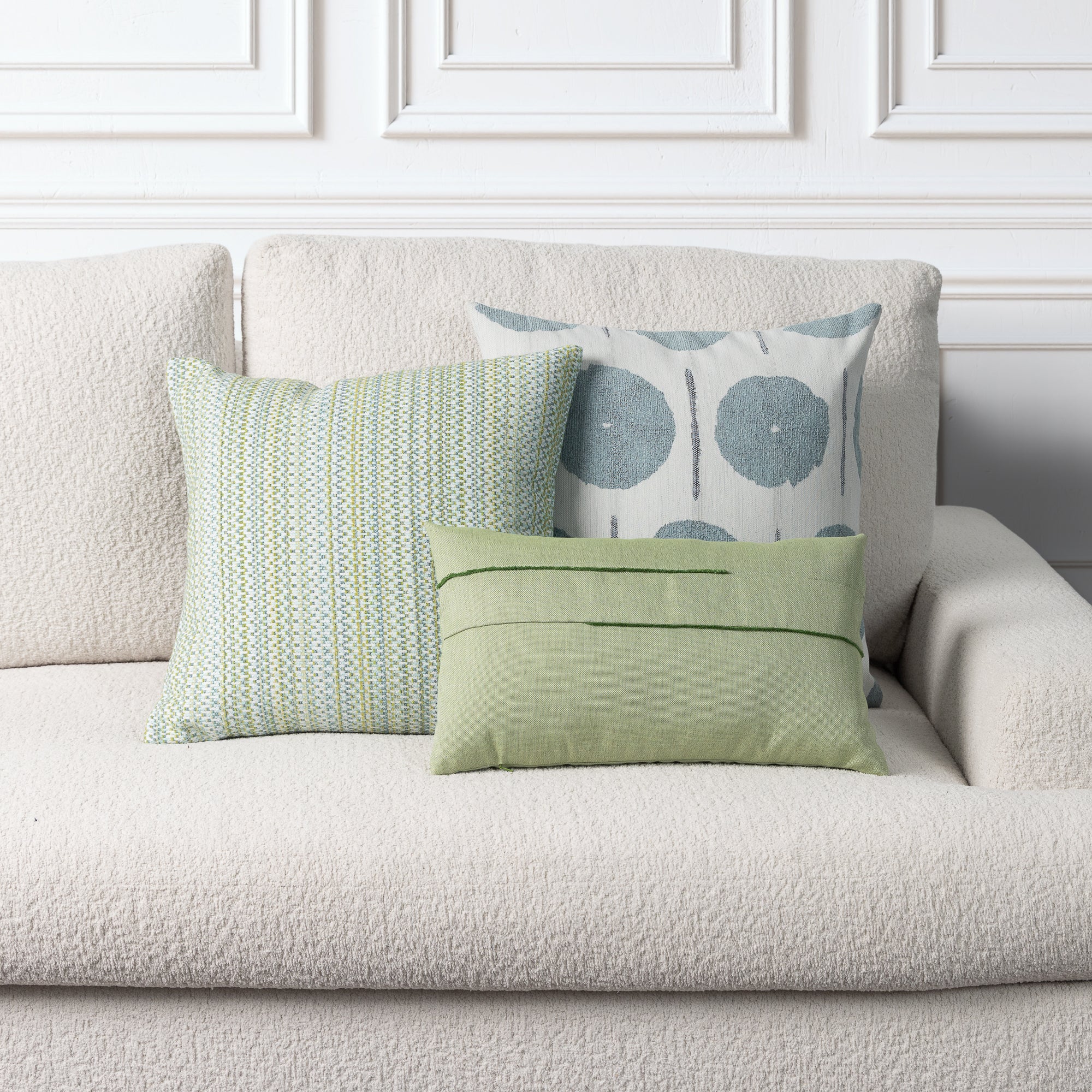 Three decorative throw pillows styled on a textured cream bouclé sofa against a white paneled wall. The pillows include a sage green lumbar pillow with horizontal stitching, a striped mint green and white pillow, and a white pillow with blue-gray circular patterns.