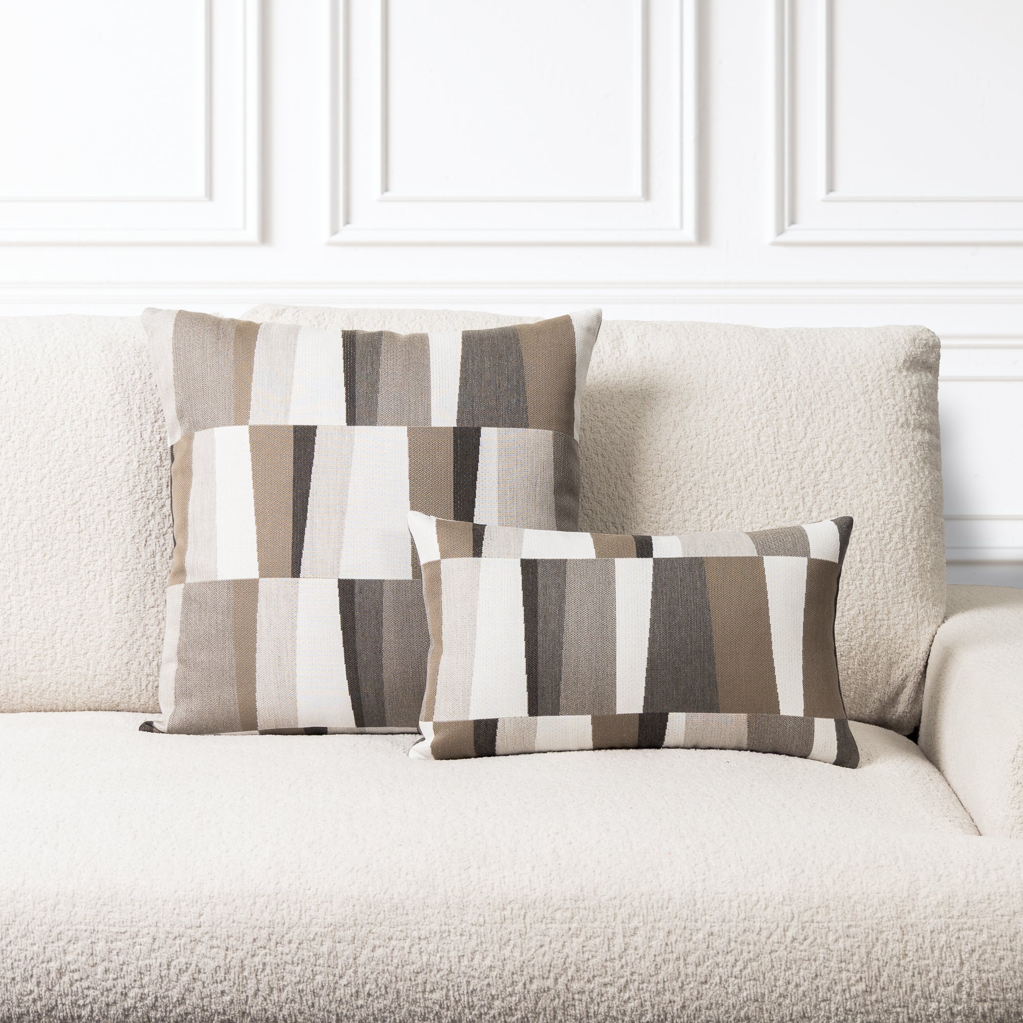 Two rectangular throw pillows with a geometric stripe pattern in beige, brown, gray, and white, styled on a textured cream sofa against a white paneled wall.