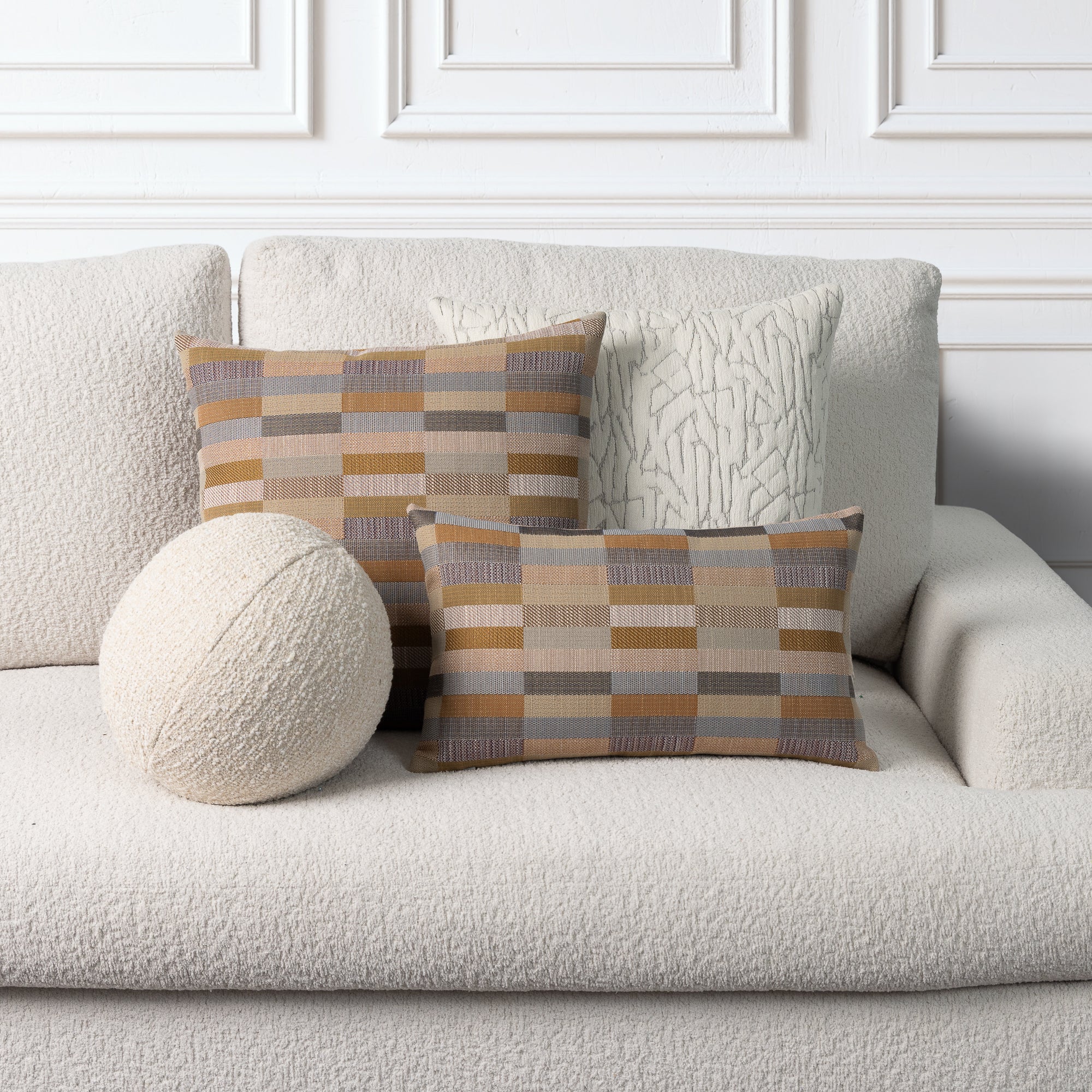 Two rectangular throw pillows with a geometric checkered pattern in beige, brown, and gray, displayed on a textured off-white sofa alongside a round textured cream pillow.