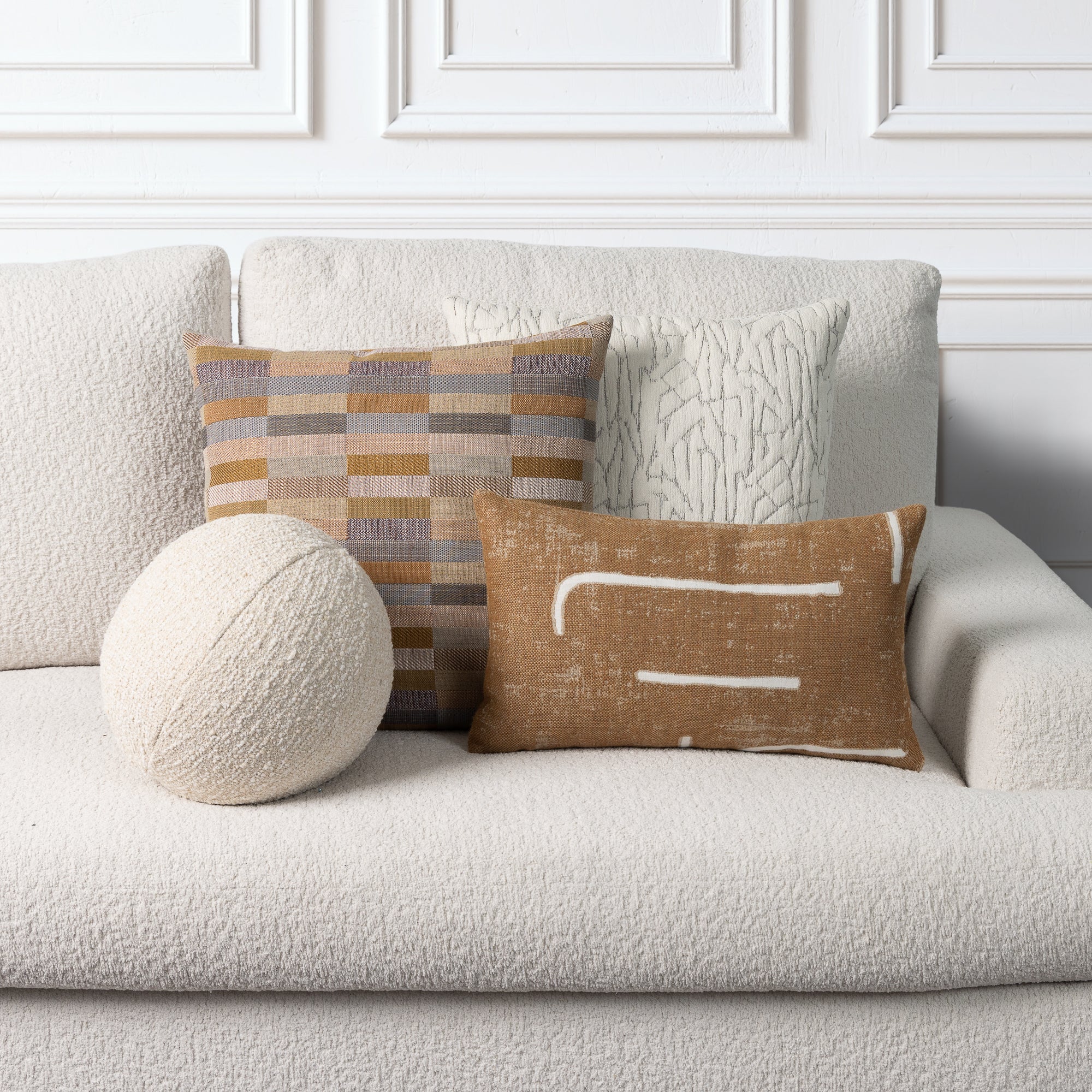 Three decorative throw pillows styled on a textured cream boucle sofa against a white paneled wall. The set includes a round cream knit pillow, a rectangular striped pillow in beige, brown, and gray, and a brown linen-texture pillow with white painted lines.