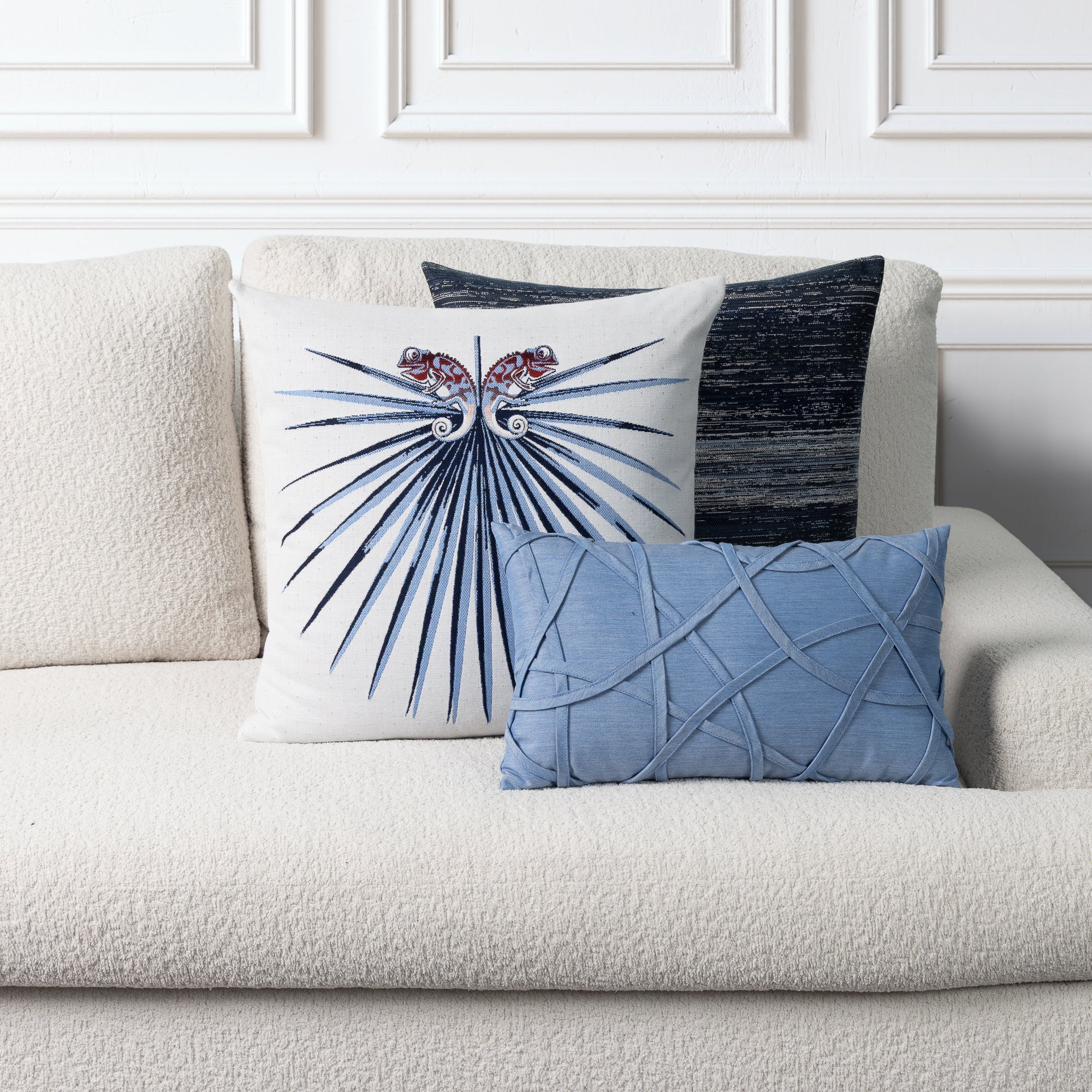 Three decorative throw pillows arranged on a cream bouclé sofa against a white paneled wall. The front pillow is white with a blue fanburst motif and red floral accents, the middle pillow is dark blue-gray with a textured vertical stripe, and the front pillow in front is light blue with a raised lattice pattern.