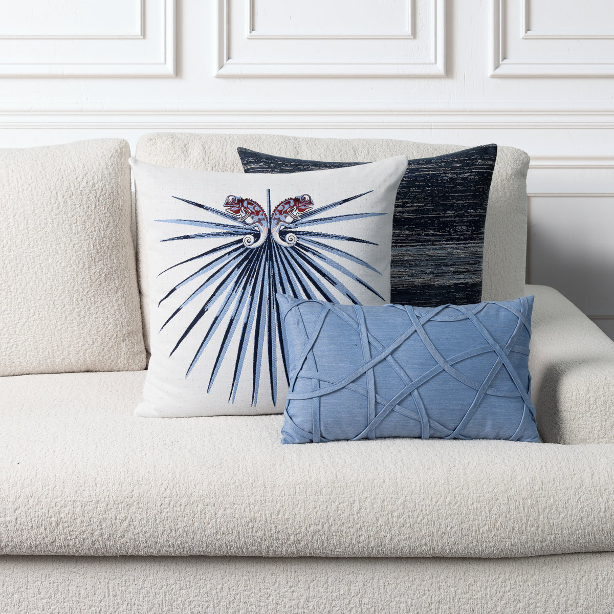 Three decorative throw pillows arranged on a cream bouclé sofa against a white paneled wall. The front pillow is white with a blue fanburst motif and red floral accents, the middle pillow is dark blue-gray with a textured vertical stripe, and the front pillow in front is light blue with a raised lattice pattern.