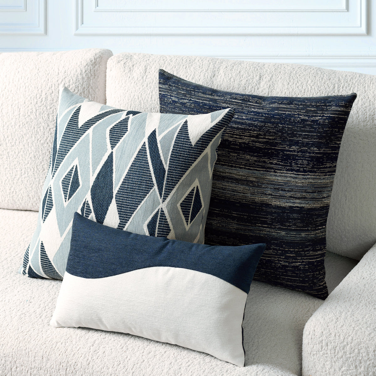 Three decorative throw pillows arranged on a cream boucle sofa. The pillows include a square pillow with a geometric pattern in navy, gray, and off-white, a textured dark navy square pillow, and a smaller rectangular pillow with a navy top half and off-white bottom half.