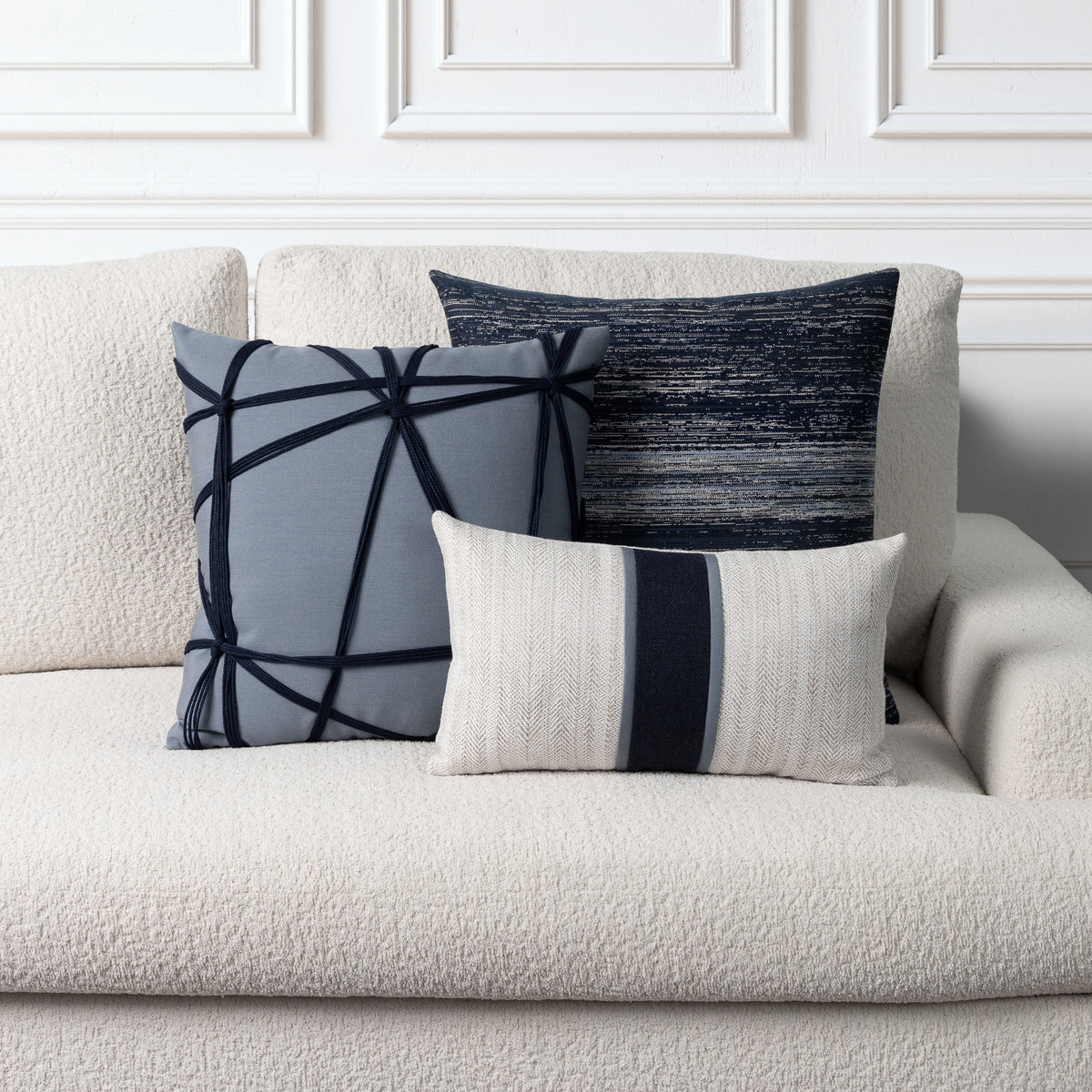 Three decorative throw pillows in shades of gray, navy, and cream are arranged on a textured cream boucle sofa against a white paneled wall.