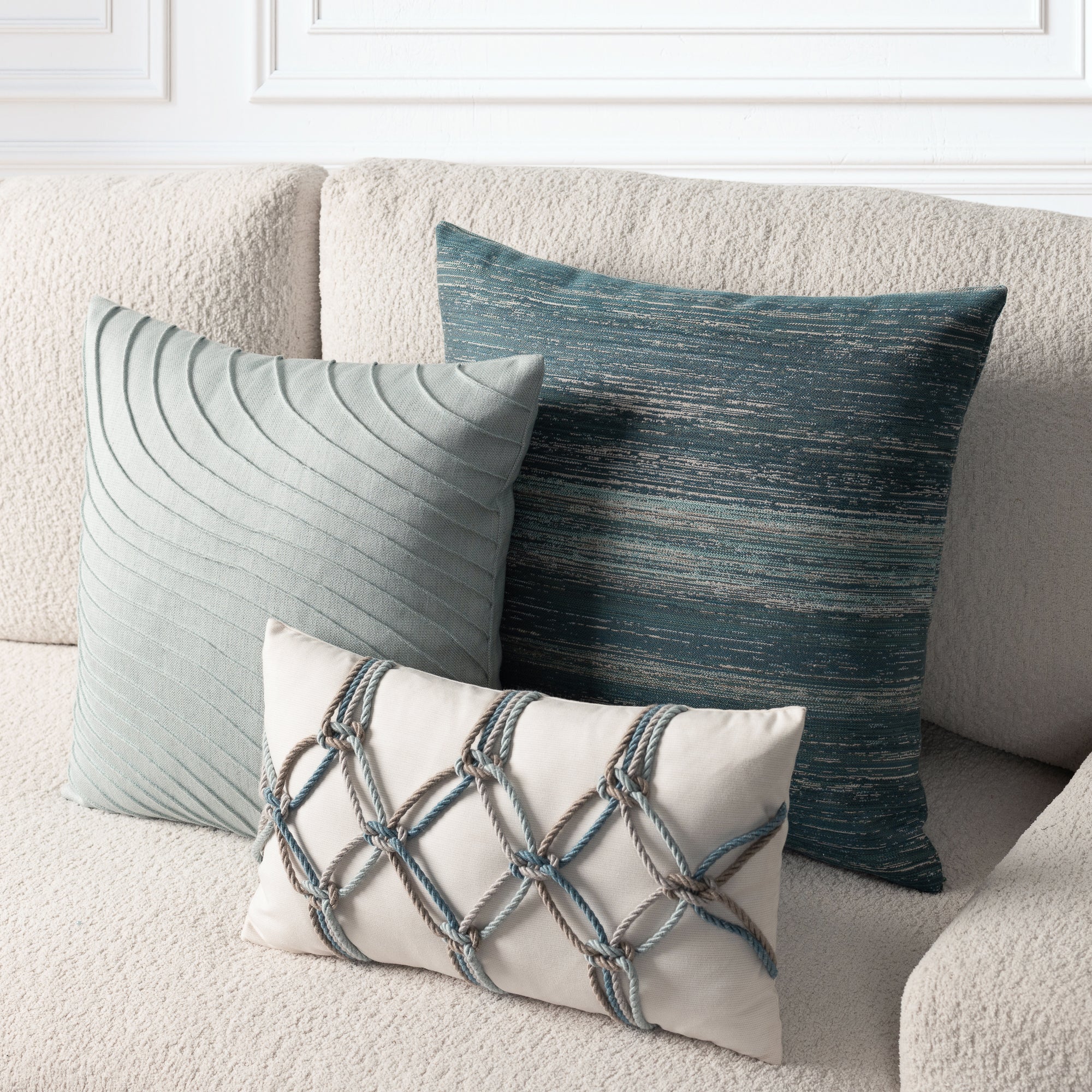 Three decorative throw pillows arranged on a textured cream sofa: a large slate blue-green pillow with a textured horizontal pattern, a smaller pale blue-green pillow with curved pleats, and a small cream pillow with a blue-gray rope lattice design.