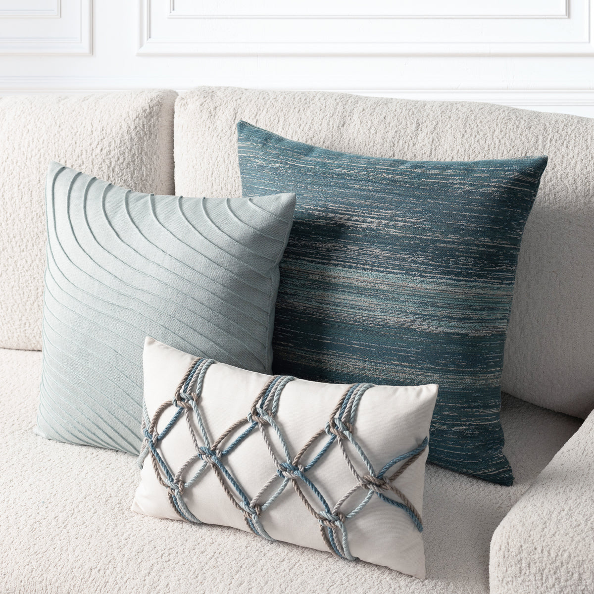 Three decorative throw pillows arranged on a textured cream sofa: a large slate blue-green pillow with a textured horizontal pattern, a smaller pale blue-green pillow with curved pleats, and a small cream pillow with a blue-gray rope lattice design.