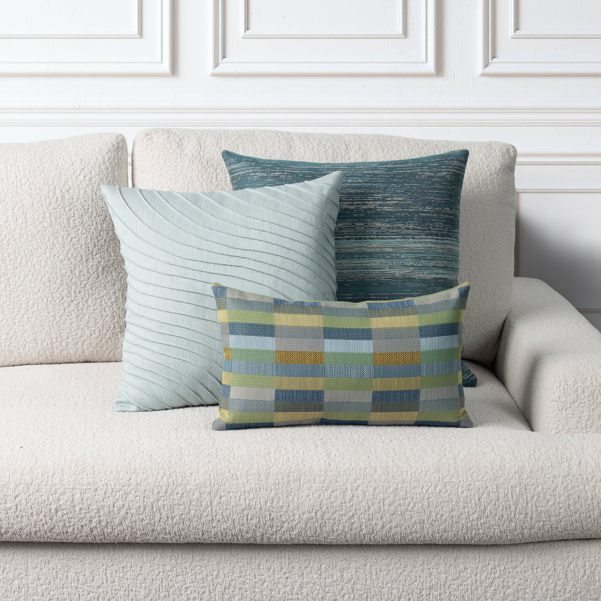 Three decorative throw pillows arranged on a textured cream sofa against a white paneled wall. The front pillow is a small rectangular plaid cushion in blue, green, yellow, and gray.