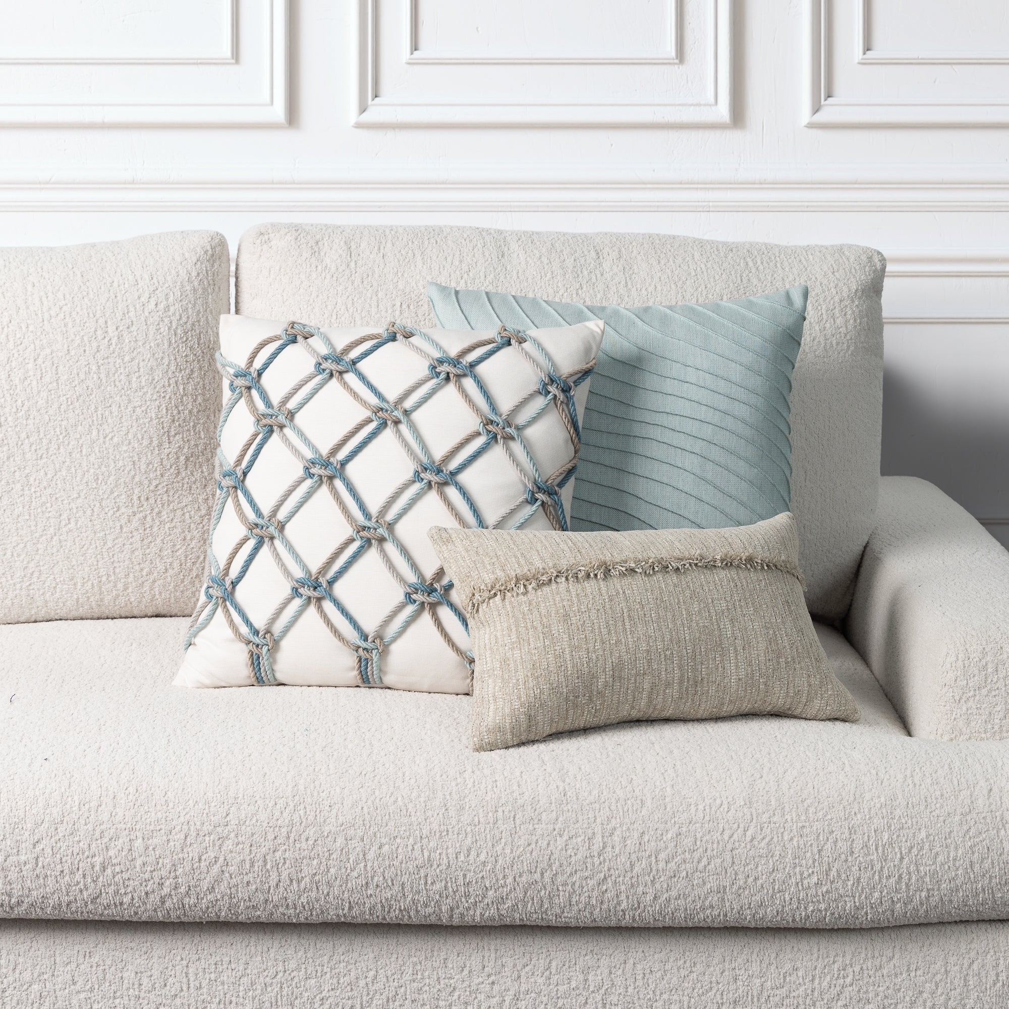 Three decorative throw pillows styled on a textured cream boucle sofa against a white paneled wall. The pillows include a square cream pillow with a blue and gray rope lattice pattern, a light blue-green ribbed square pillow, and a small beige fringed lumbar pillow.