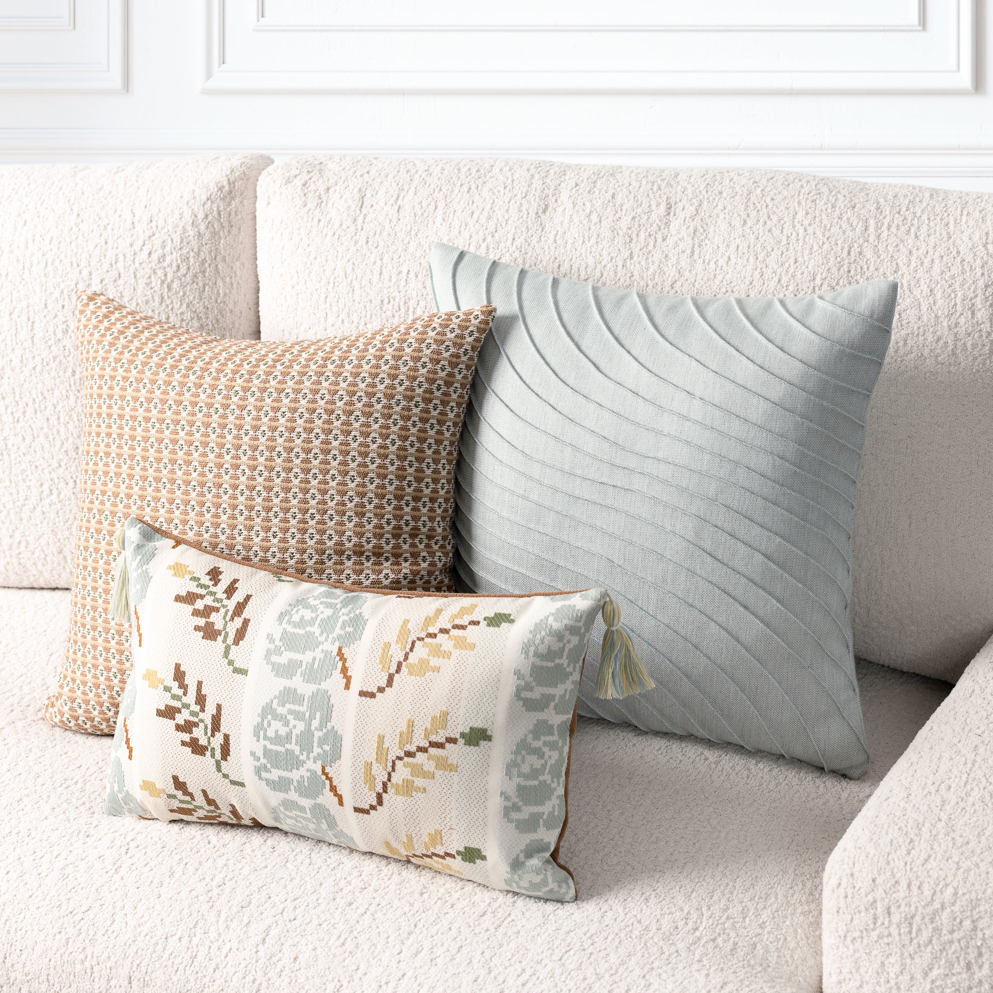 Three decorative throw pillows arranged on a textured cream sofa in a bright, modern room. The pillows include a large sage green pillow with a wavy quilted pattern, a tan and brown geometric patterned pillow, and a smaller cream pillow with a muted floral and leaf pattern in sage green, brown, and mustard.