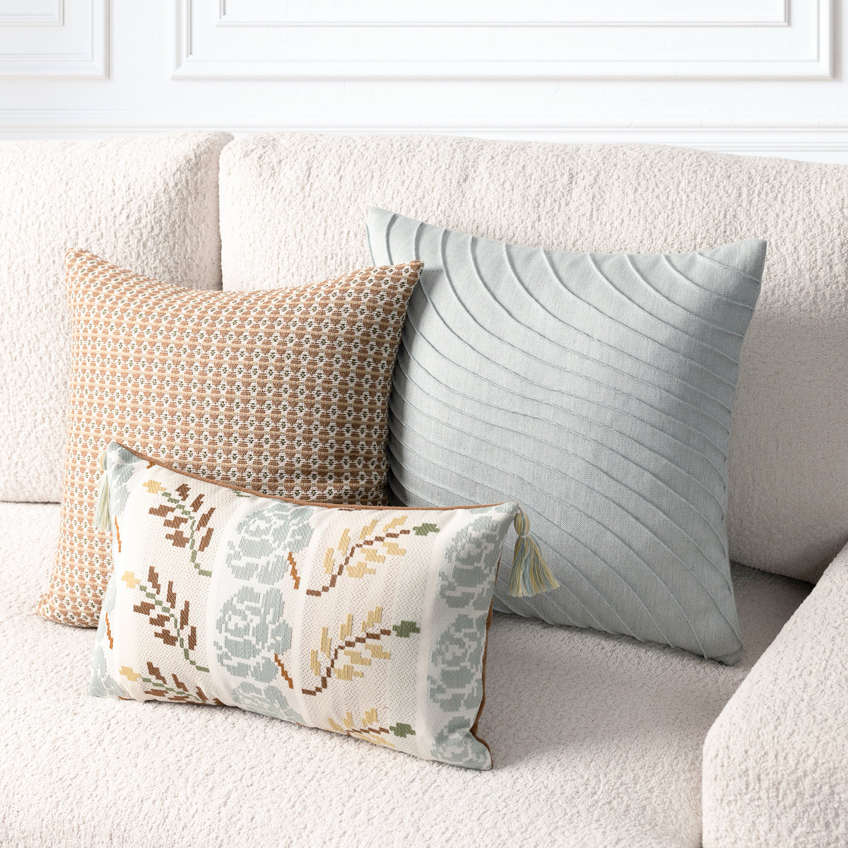 Three decorative throw pillows arranged on a textured cream sofa in a bright, modern room. The pillows include a large sage green pillow with a wavy quilted pattern, a tan and brown geometric patterned pillow, and a smaller cream pillow with a muted floral and leaf pattern in sage green, brown, and mustard.