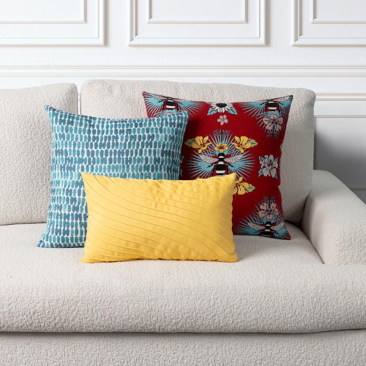 Three decorative throw pillows arranged on a light beige boucle sofa against a white paneled wall. The pillows include a turquoise and white patterned square pillow, a red floral pillow with black and yellow details, and a yellow pleated rectangular lumbar pillow.
