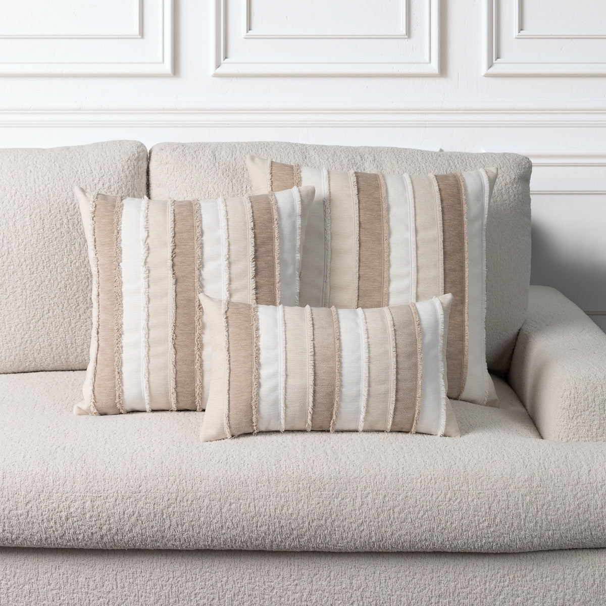Three striped throw pillows in beige, cream, and taupe tones arranged on a textured light beige sofa against a white paneled wall.